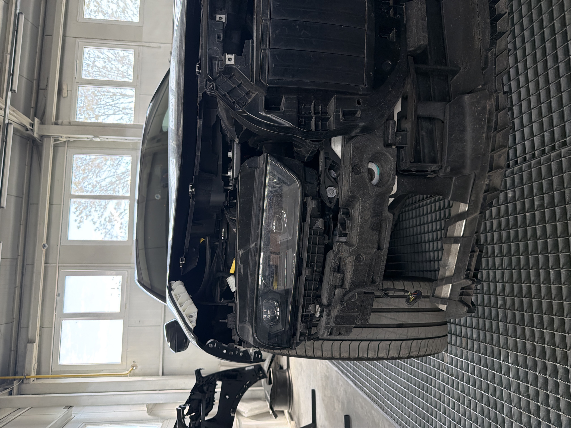 The image shows the front of a car undergoing repair or maintenance in a garage or workshop. The front bumper and some other parts have been removed, exposing the headlight, the supporting structure and some of the car's internal components. A tire is visible. The setting appears to be an indoor location with natural light coming through windows along the left side. The floor is a metal grid. I do not have sufficient information to determine the specific location in Burgas, Bulgaria.