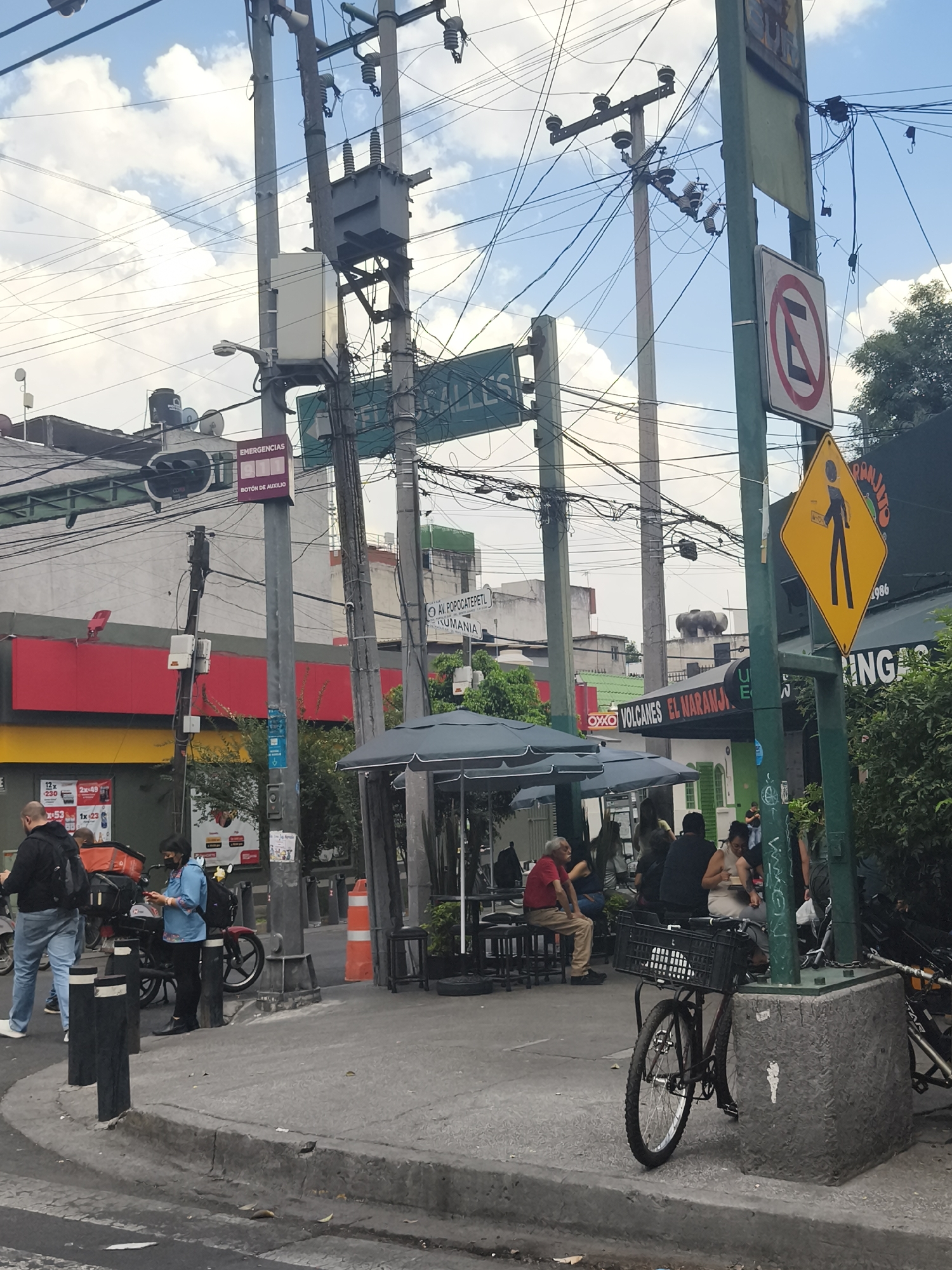 The image shows a street scene in Mexico City, Mexico. Several people are present, including those gathered at an outdoor cafe and individuals interacting near a delivery motorbike. The cafe has tables with umbrellas and is located beneath a storefront sign that reads "OXXO VOLCANES EL NARANJO." Various street signs are visible, including a green sign indicating "CALLES," an "Emergencias Botón de Auxilio" sign, a "No Parking" sign, and a yellow pedestrian crossing sign. A street name is also visible indicating "AV POPOCATEPETL ROMANIA." A bicycle with a basket leans against a street fixture. Utility poles laden with wires dominate the skyline. A building with a red and yellow facade is also in view.