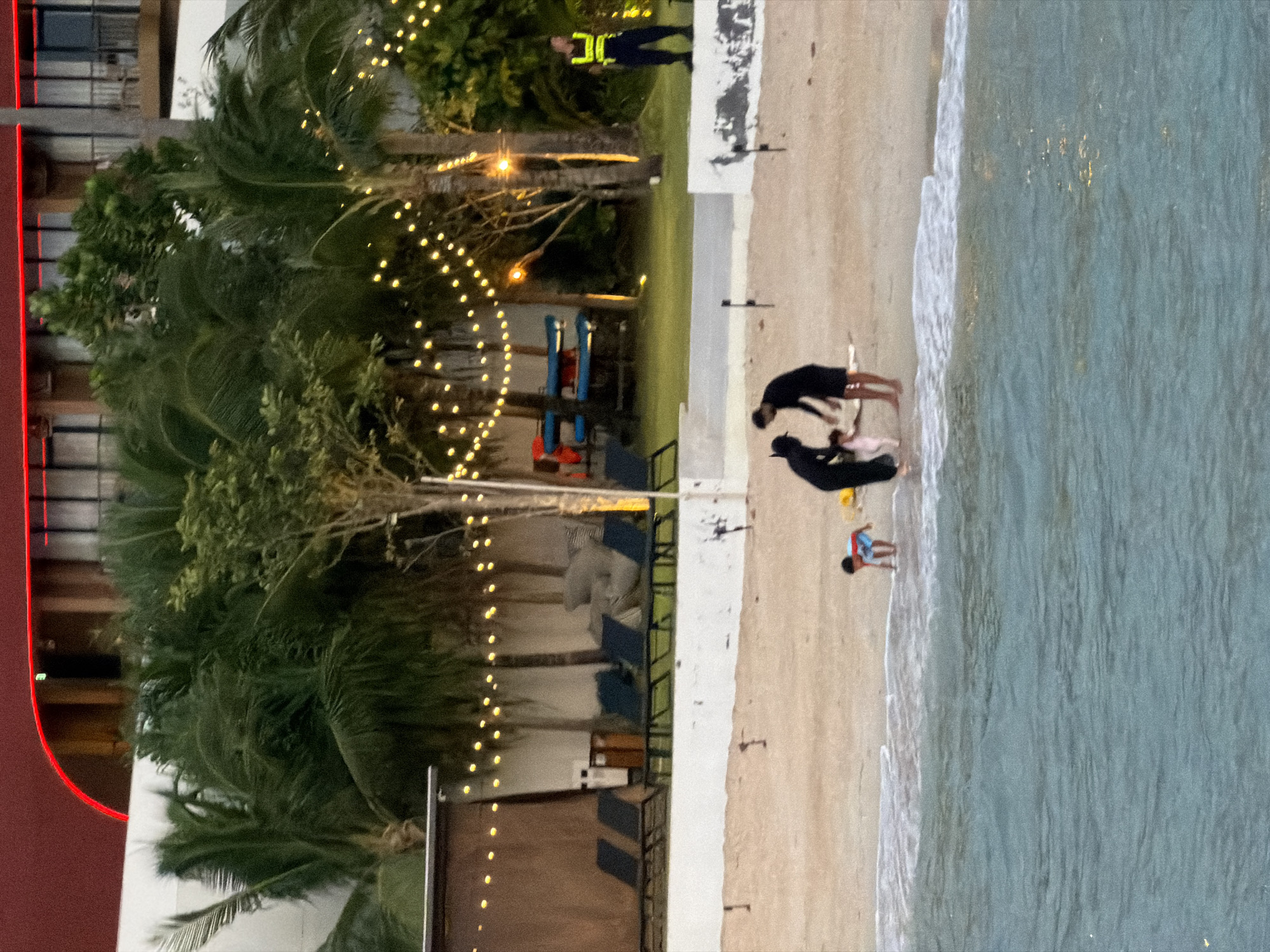 The image depicts a beach scene in Ko Samui District, Thailand, during what appears to be twilight or evening. In the foreground, the turquoise water of the sea meets the sandy shore with gentle waves.
On the beach, two adults, both dressed in dark clothing, are interacting with a young child. One adult is bent over, seemingly attending to the child who is also near the water's edge. Another child in a blue and red striped shirt is further down the beach, closer to the water, appearing to crouch or play.
In the background, a landscaped area features palm trees and other tropical foliage. String lights are illuminated, creating a festive atmosphere. Behind the greenery, a modern building with balconies and a distinctive red curved roof is visible. A person in a high-visibility vest stands on a raised concrete platform, possibly a lifeguard or security personnel. Lounge chairs are arranged on the sand near a covered structure. The scene suggests a resort or hotel setting.