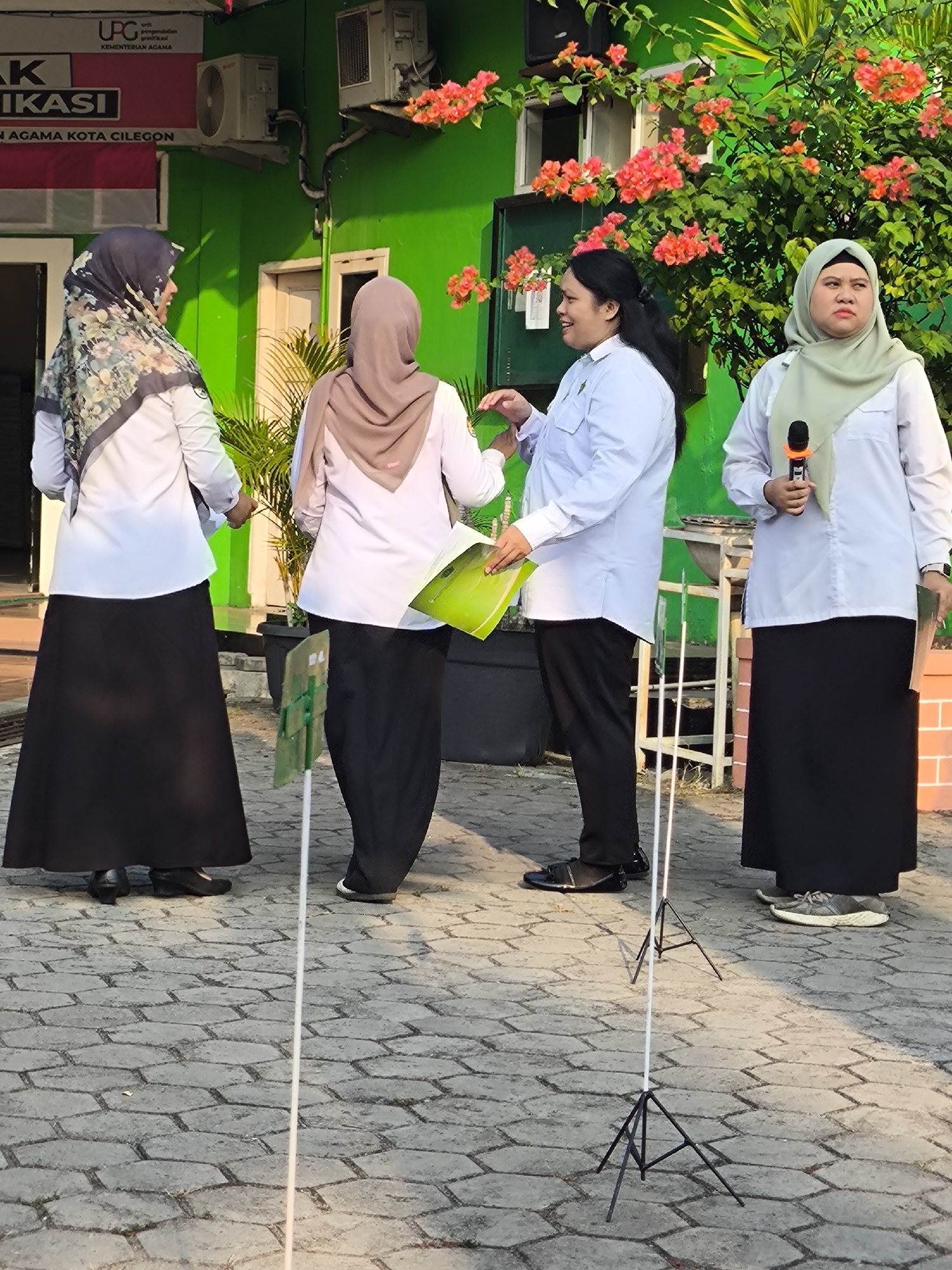 Four individuals, two men and two women, are standing in front of a bright green building in Cilegon, Indonesia. The building has an overhang with signage that reads "UPT unit pengendalikan karir", "KEMENTERIAN AGAMA", and "NK AGAMA KOTA CILEGON". The woman on the far left is wearing a patterned hijab and a white long-sleeved shirt with a black long skirt. The second person from the left is wearing a brown hijab and a white long-sleeved shirt with a black long skirt. The third person from the left, a man, is wearing a white long-sleeved shirt and black pants. He is holding a green folder. The woman on the far right is wearing a light green hijab and a white long-sleeved shirt with a black long skirt. She is holding a microphone. The ground is paved with hexagonal bricks. There are some potted plants and flowering bushes in the background.