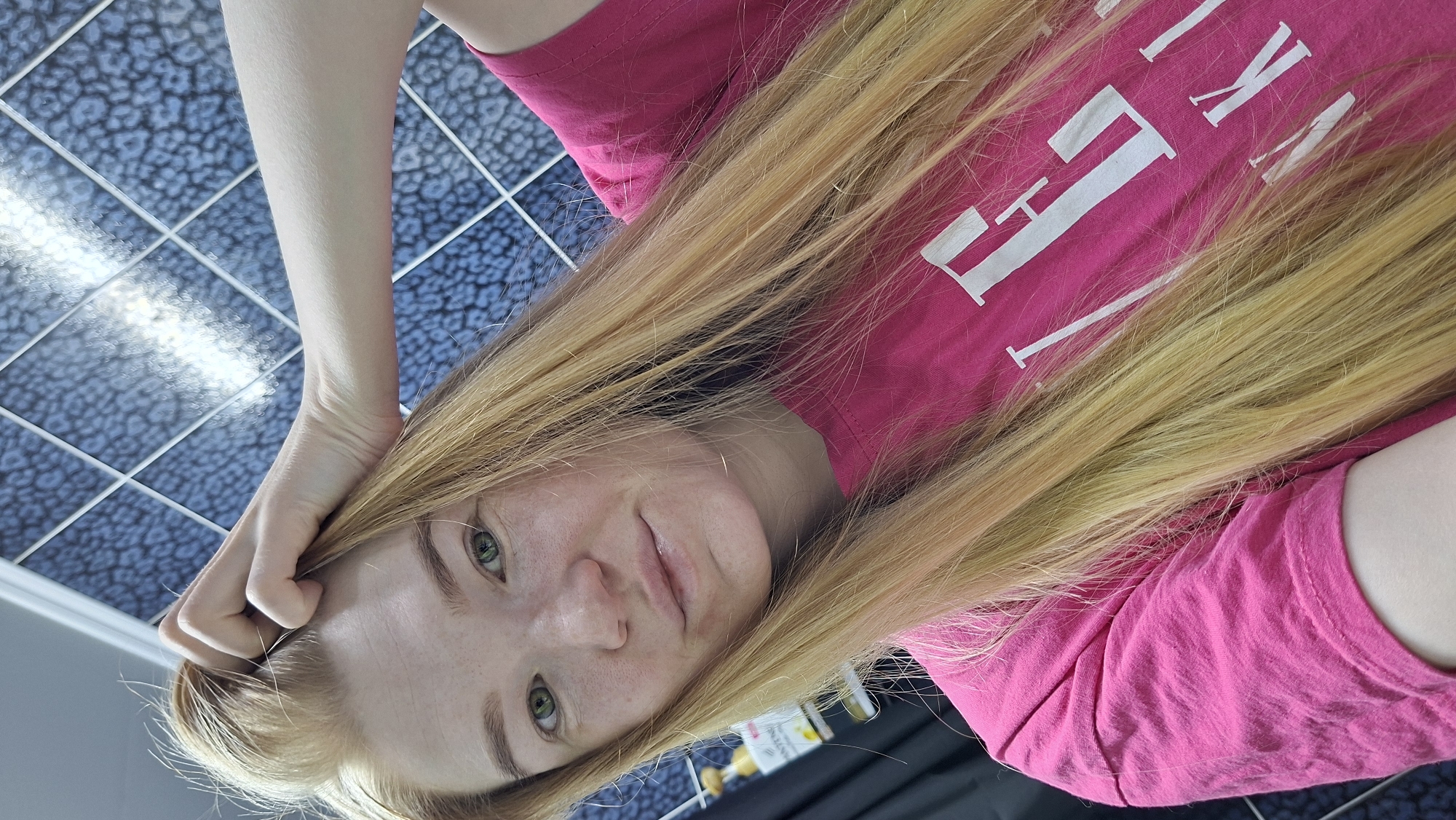 A woman with long blonde hair and green eyes is positioned in front of a blue and white tiled floor. She is wearing a pink t-shirt with white lettering. Her left hand is touching her hair. The location is not identifiable as Chaikovsky, Russia based on the visual information provided.