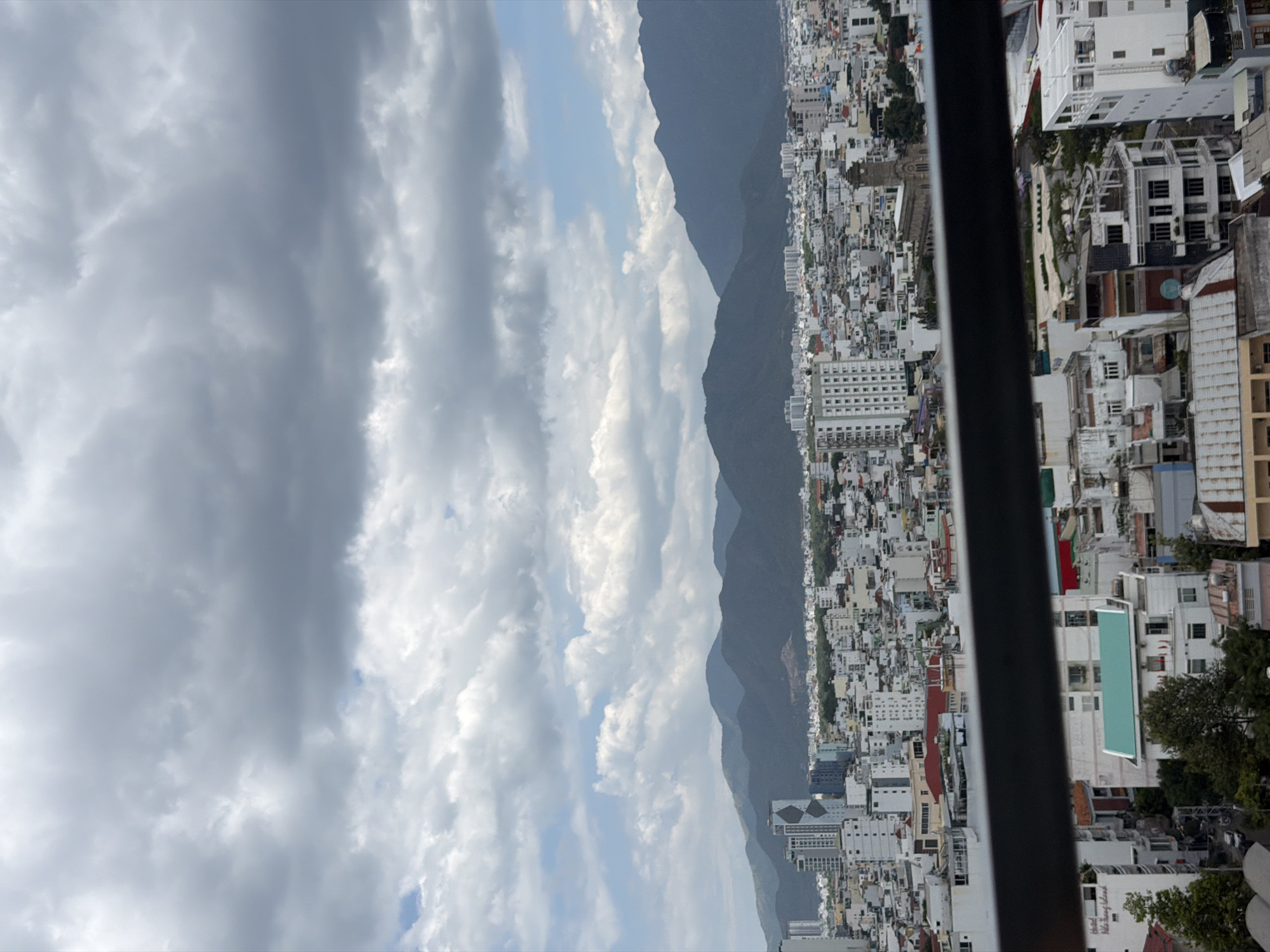 The image captures a panoramic view of Nha Trang, Vietnam, from an elevated vantage point. In the foreground, a dark railing partially obscures the lower part of the scene, which consists of a dense urban landscape. Numerous buildings, varying in height and architectural style, are tightly packed together. Many of these structures are residential or commercial, displaying white or light-colored facades, tiled roofs in shades of red and green, and balconies. Some taller, modern buildings with glass windows stand out amongst the lower-rise structures.
Further back, the city stretches towards a range of dark, mountainous hills that form the backdrop. The mountains are covered in vegetation and appear to recede into the distance. The sky above is a mix of dramatic, gray storm clouds and patches of bright blue sky with scattered white clouds, suggesting a dynamic weather system. The overall impression is one of a bustling coastal city nestled between mountains and the sea. No individuals are clearly discernible in the image, and no specific actions or interactions are captured. The focus is on the urban sprawl and the natural landscape.