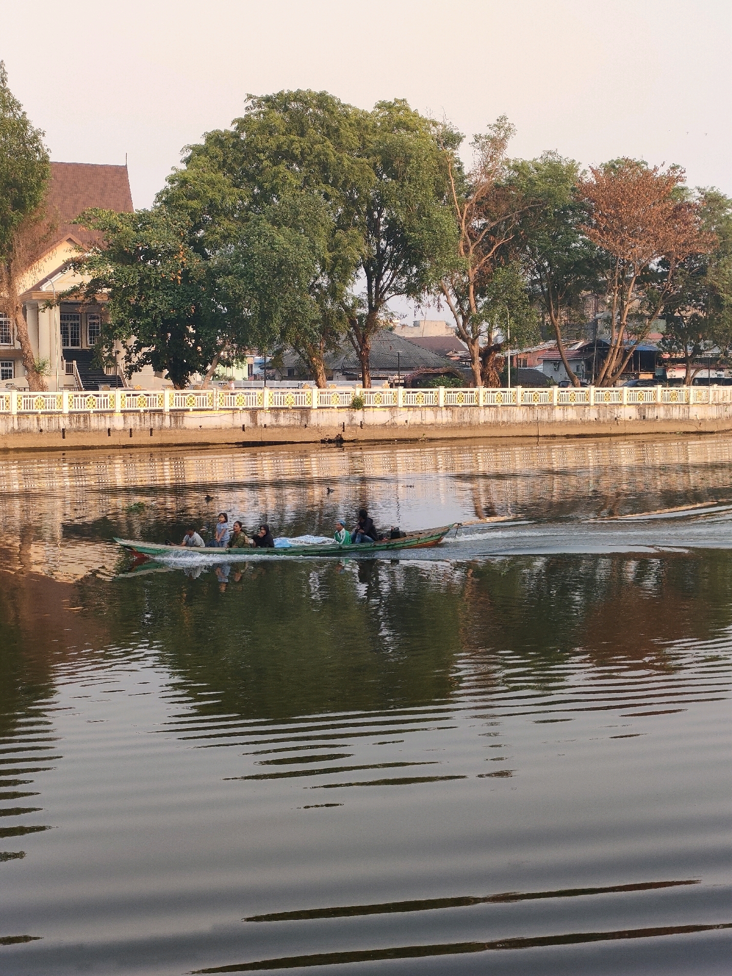 The image depicts a boat carrying several people on a river in Banjarmasin, Indonesia. The boat, a long and narrow vessel, is moving through the water, creating a wake. Onboard, there are at least six individuals, including men, women, and children. Some are seated, while others appear to be standing or adjusting their positions. The river itself is calm, with reflections of the surrounding environment visible on its surface.
In the background, the riverbank is lined with a decorative white fence with golden accents. Behind the fence, a large building with a brown roof and white columns is visible, likely a public building or a large residence. Mature trees with lush green foliage stand tall along the riverbank, some with autumnal brown leaves, suggesting a seasonal change or varied species. Further in the background, other buildings and structures can be seen, indicative of a populated urban or semi-urban area. The sky above is a pale, hazy color, characteristic of a warm day or possibly a slight atmospheric haze. The overall scene suggests a tranquil moment on the water, with daily life unfolding against a backdrop of traditional Indonesian architecture and natural greenery.