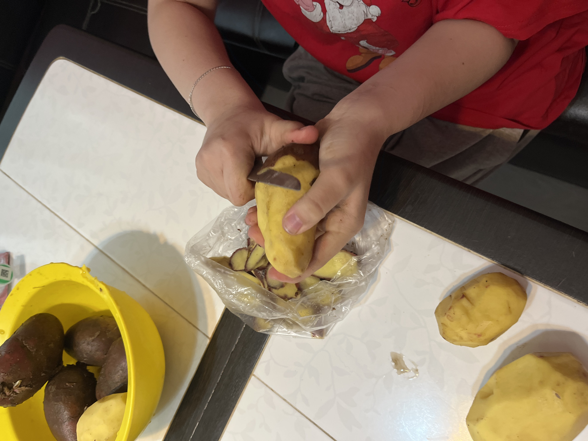 The image captures a top-down view of a person peeling potatoes indoors, likely in a kitchen or dining area in Semyonov, Russia. The individual, whose face is not visible, wears a red t-shirt with a white cartoon character print (resembling Santa Claus) and grey pants. A delicate silver chain bracelet adorns their left wrist.
The primary activity shows the person's hands holding a yellow potato and a small knife, actively peeling the skin. The potato peels are directed into a clear plastic bag positioned below, which already contains several other peels.
To the left of the scene, a vibrant yellow plastic bowl holds a collection of potatoes, predominantly dark purple-skinned varieties, along with at least one yellow-skinned potato. A small white sticker with a partial QR code is visible on the side of the yellow bowl. On the right side of the white table, two more yellow potatoes are present; one is mostly unpeeled, and the other is partially peeled.
The work surface is a white table with a subtle, light-colored floral or leaf pattern. Adjacent to the white table is a dark brown or black surface. The overall impression is one of domestic food preparation. No specific time of day or emotions are discernible.