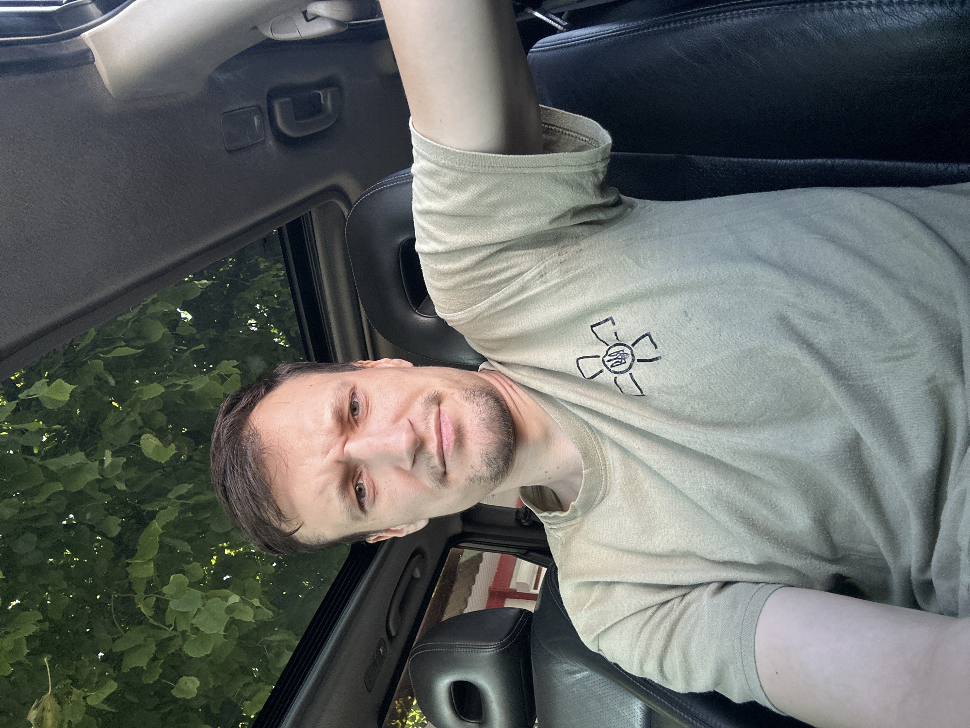 A selfie is taken from inside a car, looking forward. The car has a sunroof, through which green leaves and sunlight are visible, suggesting an outdoor, possibly wooded or tree-lined location. The man in the photo has short brown hair, a light beard, and is wearing a light-colored t-shirt with a black emblem of a stylized flower containing a trident. He is seated in the driver's seat, with the passenger seat and headrest visible to his left. The interior of the car appears to be black leather. The man's left arm is resting on the center console. The background visible through the side window suggests buildings with red elements. The scene is consistent with being parked somewhere within the city of Kamianets-Podilskyi, Ukraine, likely in an area with trees and structures.