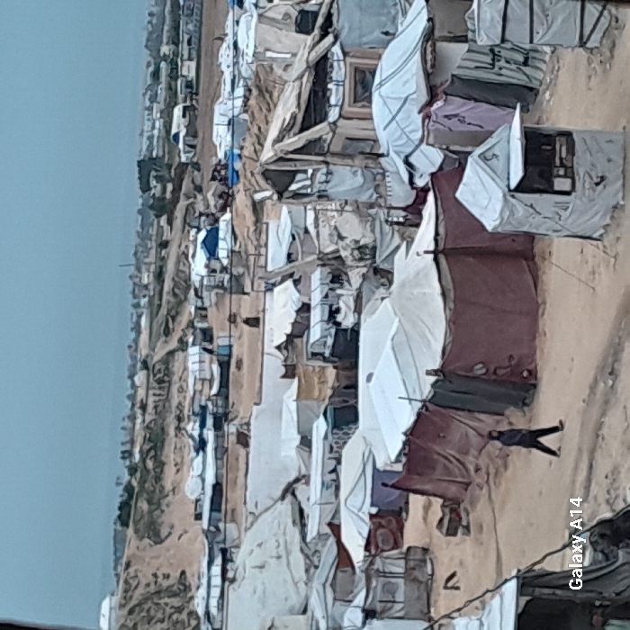 The image depicts a dense makeshift settlement, possibly a refugee camp or slum. Structures appear hastily constructed with various materials like fabric, metal sheets, and wood. Tents and shelters crowd together on barren land. A single person walks through the camp’s narrow pathways. The horizon shows more buildings and vegetation. The overall impression is one of poverty, displacement, and hardship.