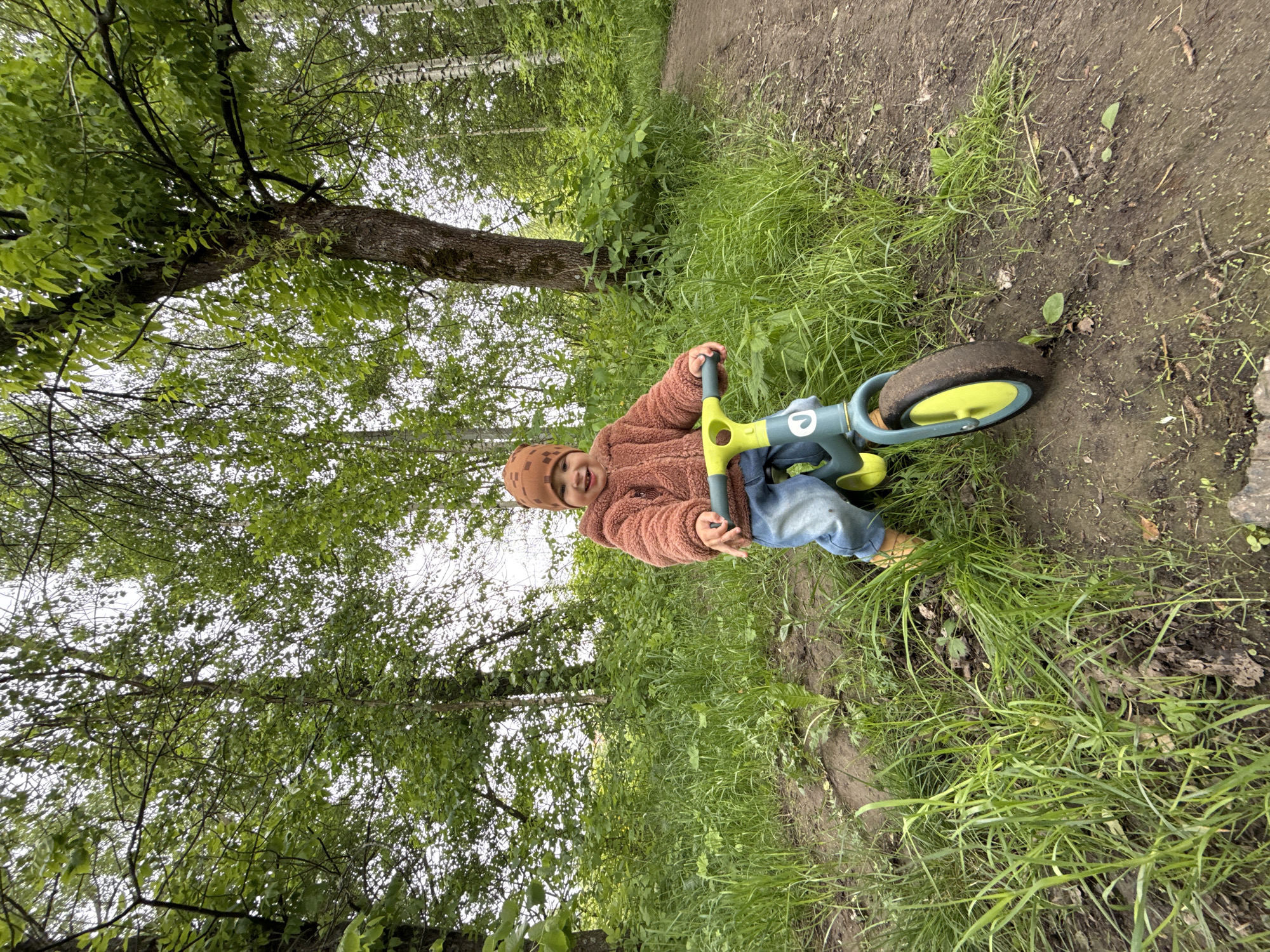 A young child wearing a brown hat, peach-colored jacket, and blue jeans sits on a blue and yellow balance bike on a dirt path in a wooded area in Rusanovo, Russia. Green grass and trees surround the path, which is made of bare earth. The sky is visible through the leaves.