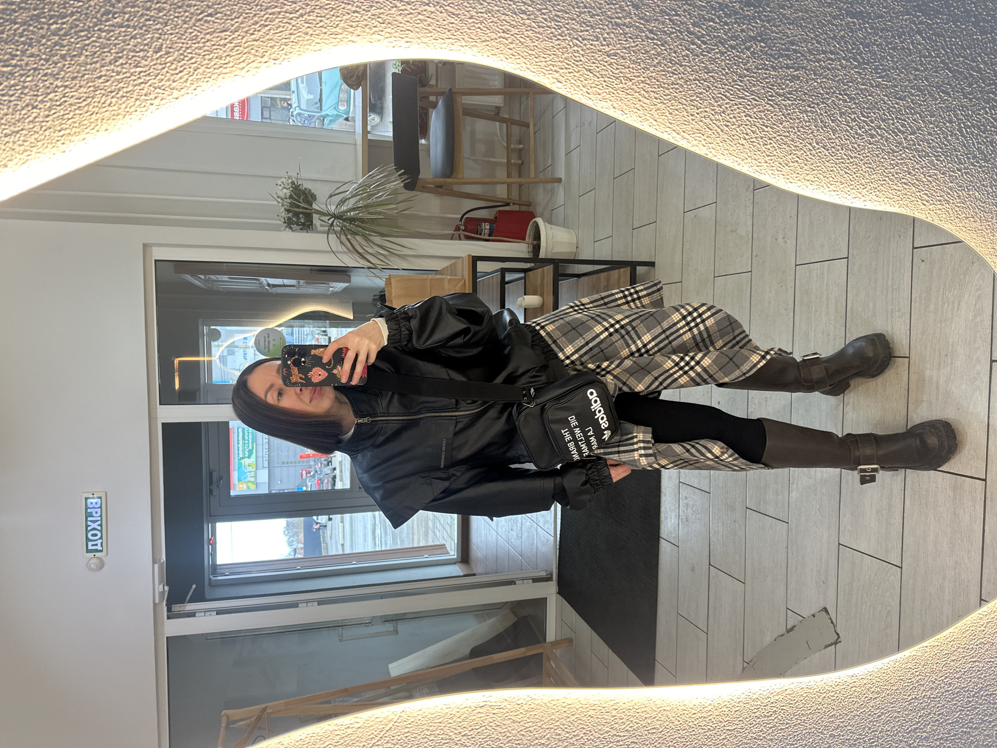A woman stands in front of a mirror in a Votkinsk, Russia establishment. She is wearing a black jacket, a plaid skirt, black leggings, and tall dark boots. She is holding a cell phone to take a selfie. The mirror reflects the interior of the establishment, including tile flooring, tables, chairs, plants, and a fire extinguisher. There is also a window with an exit sign written in Cyrillic characters.