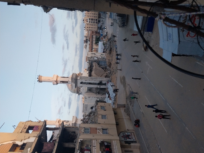 The image captures a street scene in Khan Yunis, Palestinian Territory, marked by extensive structural damage to buildings. In the mid-ground, a mosque with a light-colored dome and a tall, slender minaret stands, exhibiting visible damage to its facade and surrounding sections. To the left, a multi-story residential building shows severe destruction, with large portions of its exterior walls collapsed, revealing interior rooms and debris. Laundry is hanging from balconies on this damaged structure.
On the main asphalt street, marked with faded white lines, approximately 20-30 individuals, primarily children and young males, are present. Several small groups are actively playing soccer with a single ball in various areas of the street. Other individuals are seen walking or standing. On the left sidewalk, a group of five individuals is seated. In the foreground on the right, partially obscured by electrical wires, are remnants of what appear to be demolished or makeshift structures. Debris piles are visible on both sides of the street. The sky is partially cloudy, and the scene is illuminated by natural daylight.