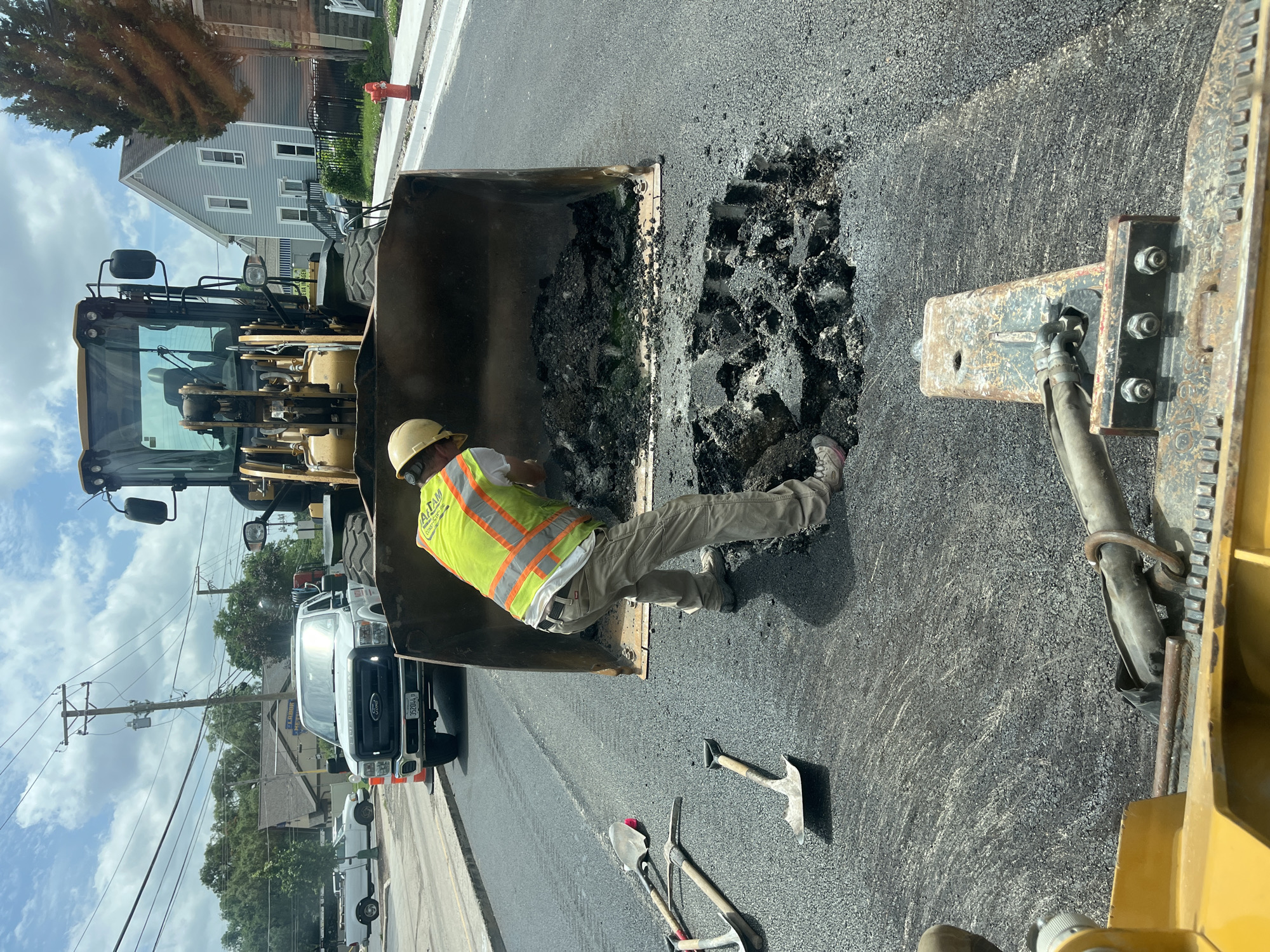 In Elgin, Illinois, USA, on a bright, partly cloudy day, a construction worker in a yellow high-visibility vest and hard hat is shoveling asphalt from the bucket of a yellow front-end loader into a large pothole on a paved street. Several shovels and a pickaxe lie on the asphalt near the pothole. A white Ford pickup truck is parked behind the loader, and houses are visible in the background, along with utility poles and wires. A red fire hydrant stands on the sidewalk. The scene appears to be a road repair operation.