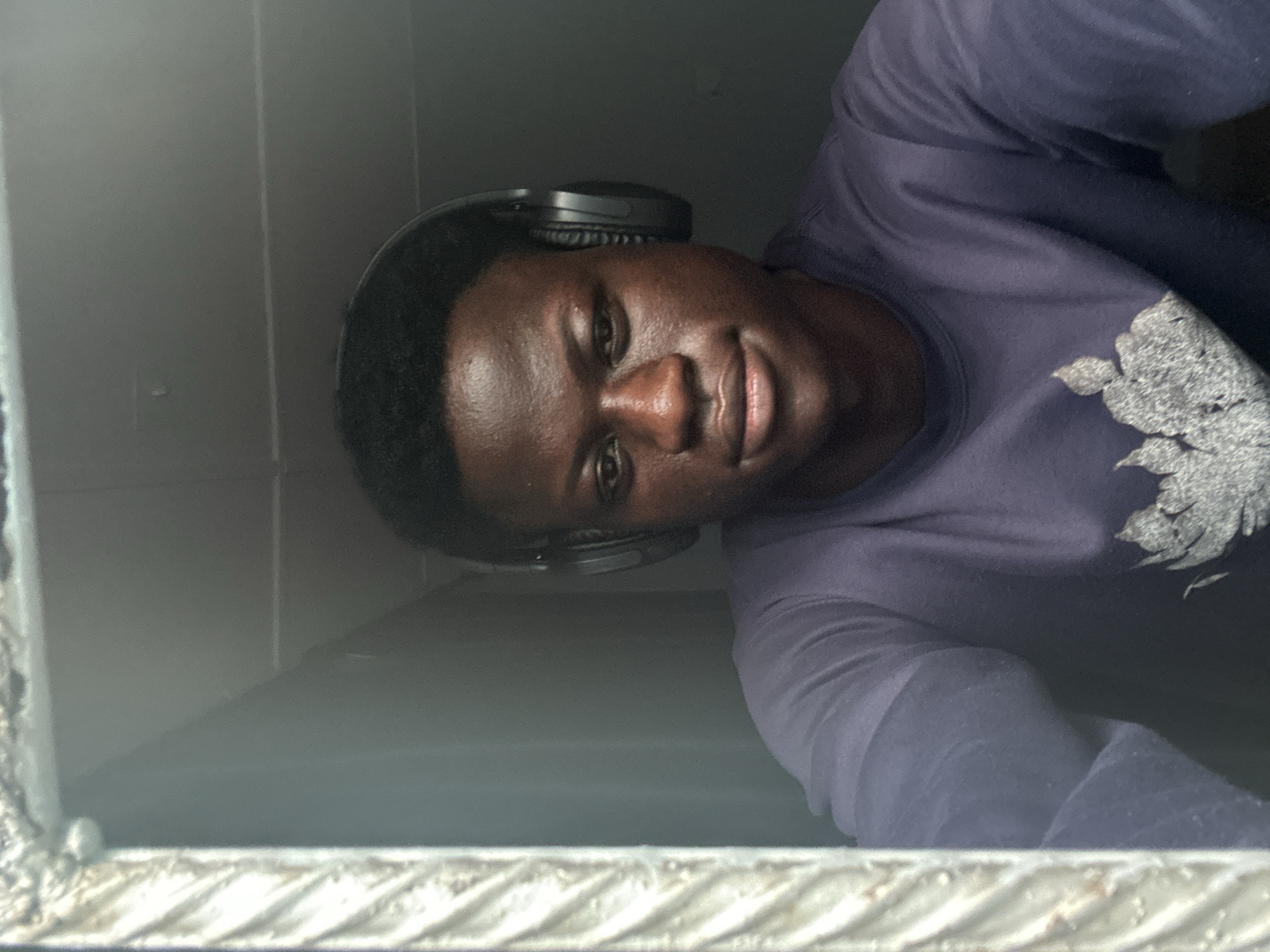 A medium shot captures a young Black male from the chest up, wearing black over-ear headphones and a dark purple long-sleeved shirt with a patterned design. He is looking directly at the camera with a neutral expression. The background appears to be an indoor space with a grey, textured ceiling and a hint of a window frame on the left, which has a light-colored, ornate metallic or wooden texture. The lighting is dim, casting shadows on the subject's face. The scene is set in Badagry, Nigeria.