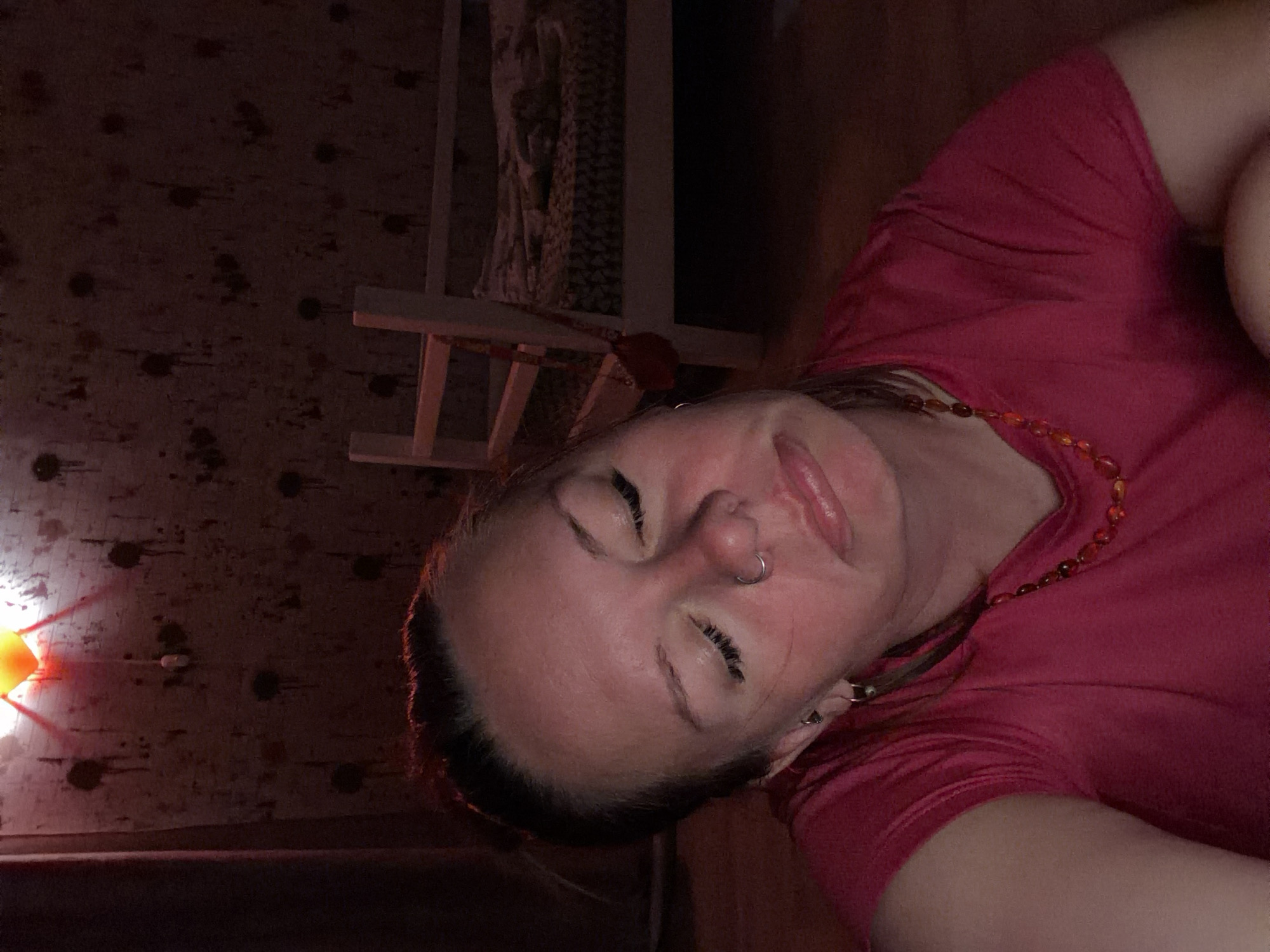 This image is a close-up, rotated selfie of a person, likely female, captured in a dimly lit indoor setting in Chaikovsky, Russia. The individual is positioned with their head to the right of the frame, eyes closed, and mouth slightly open, revealing some teeth. A silver hoop nose piercing is visible on her right nostril. Her complexion appears flushed, likely from the warm, reddish light illuminating the scene.
She is wearing a short-sleeved, fuchsia or magenta-colored top, and a necklace featuring amber-colored beads is visible around her neck. The background consists of a dark wooden wall or ceiling, textured with visible wood grain, knots, and darker stains. A prominent orange-red light source, possibly a lamp or decorative fixture, is casting a strong glow and distinct light rays from the bottom-left of the frame onto the wooden surface. Partially visible light-colored wooden furniture, resembling a chair or table leg, can be seen in the background, along with a hint of patterned fabric, possibly curtains, further back. The overall impression is one of relaxation and intimacy, set by the soft, warm lighting. There is no visible text in the media file.