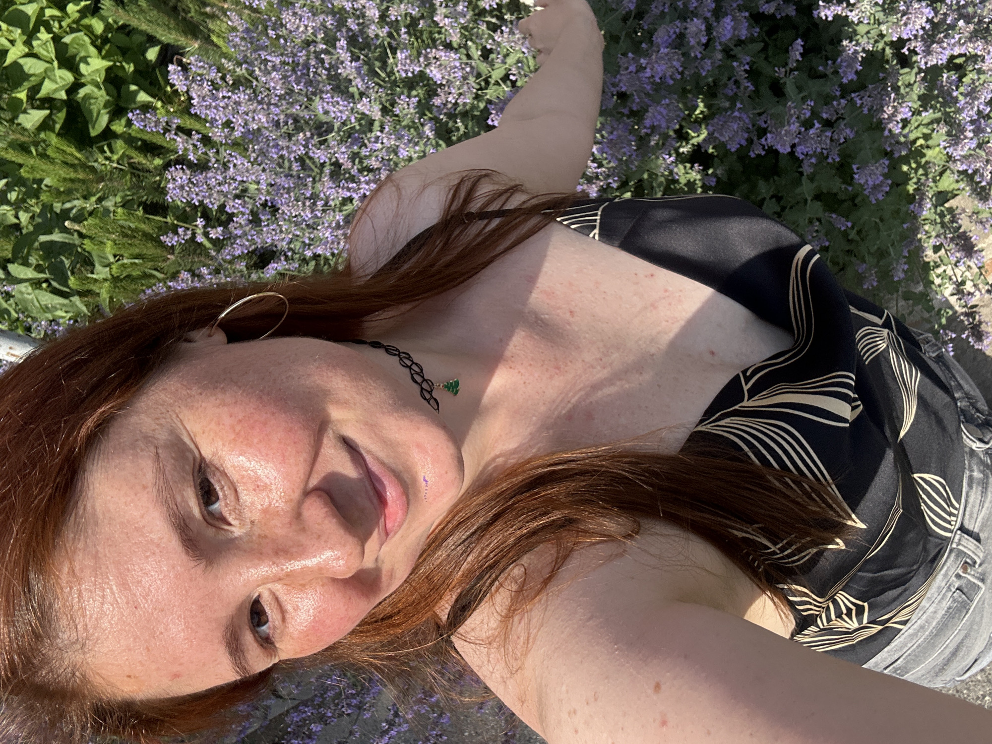 In a medium shot, a woman with auburn hair and freckles smiles into the camera while standing outdoors. She is wearing a black camisole with a gold leaf print, grey jeans, and a black choker necklace with a small green tree pendant. She has a large gold hoop earring in her left ear. The background is filled with lush green foliage and abundant purple flowers. The lighting suggests it is daytime, likely a sunny afternoon. The image does not provide any specific geographical markers that would indicate its location in Nizhny Novgorod, Russia.
