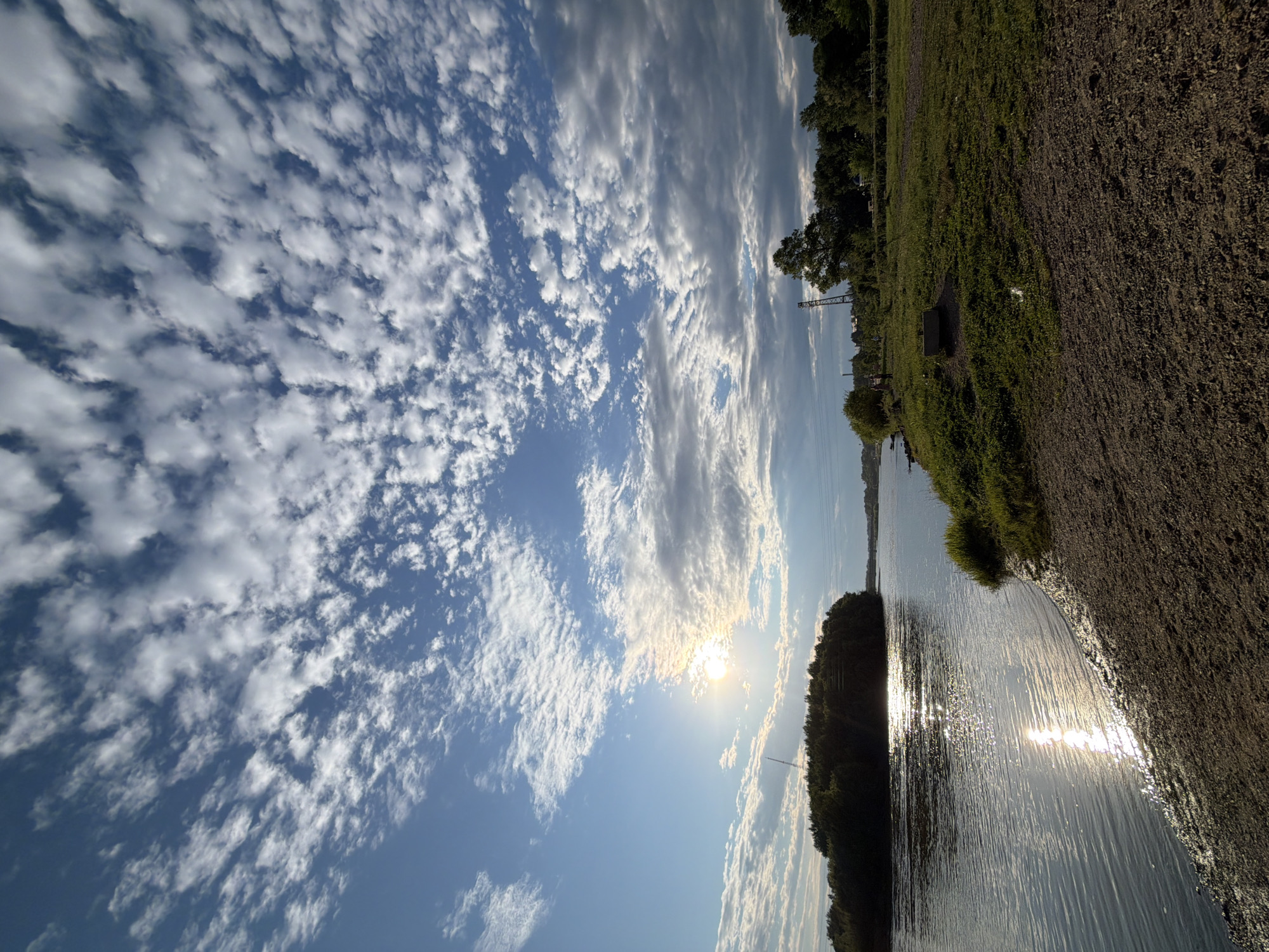 The image depicts a wide, calm river flowing through a vegetated landscape. The sun is setting or rising, casting a warm glow and reflecting brightly on the water's surface. The sky is filled with scattered clouds, some of which are illuminated by the sun. On the left side of the river, there is a dense area of trees, forming what appears to be a small island or peninsula. The right bank of the river features a grassy area with some scattered vegetation and what looks like a dirt path. In the distance, on the right bank, there are more trees and possibly some structures, including what might be a utility tower. The overall scene is natural and serene, characteristic of a riverfront area. While the image is a picturesque landscape, it does not contain any identifiable landmarks or specific features that would definitively place it within the city of Birobidzhan, Russia.