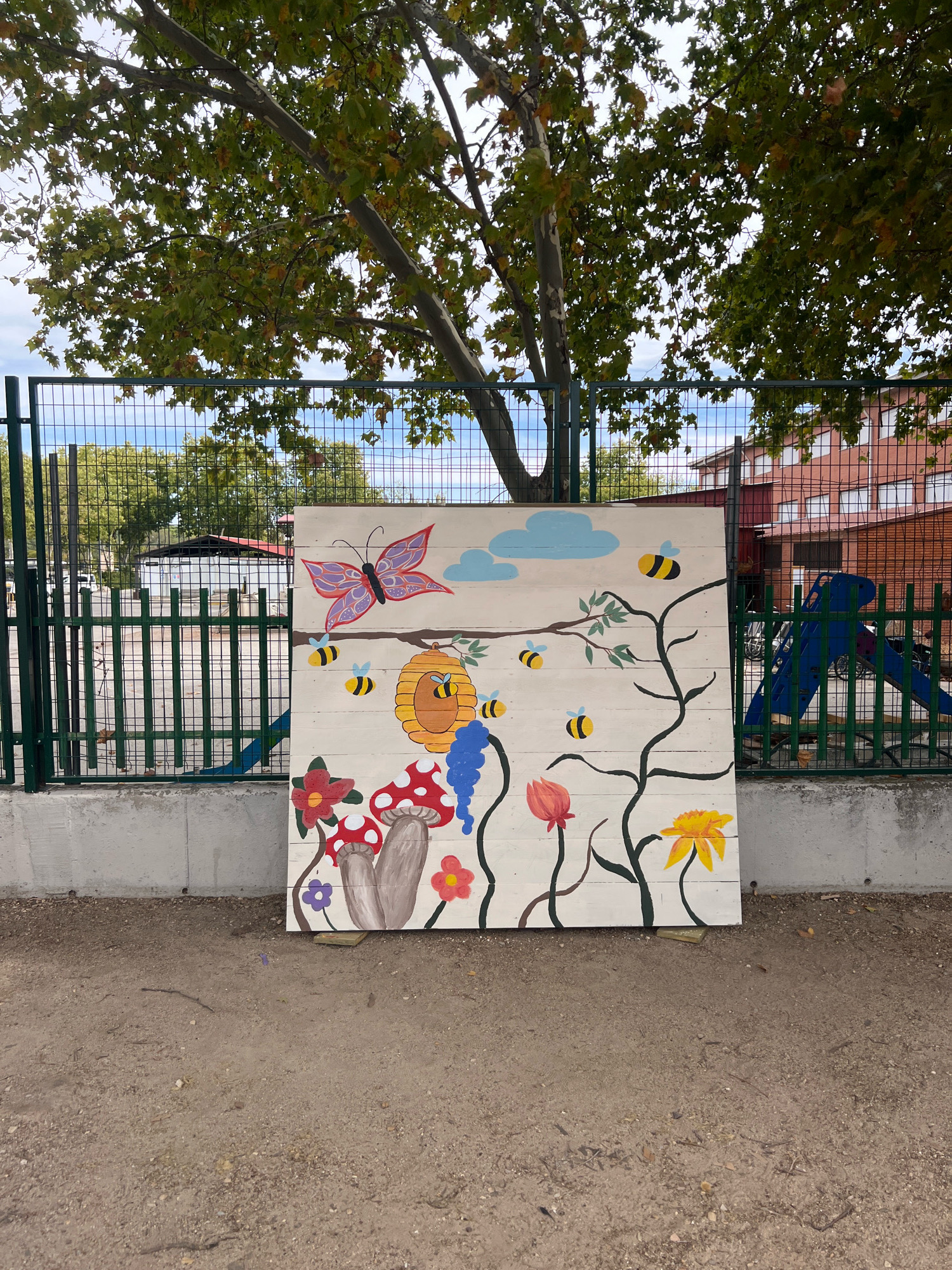 The image shows a large, rectangular wooden panel with a colorful mural painted on it. The mural depicts a whimsical scene with a large purple butterfly, several bees, a beehive, two mushrooms, and various flowers and clouds. The panel is propped up against a concrete wall in what appears to be an outdoor area. Behind the panel is a green metal fence, and beyond that, trees with green leaves and some autumn foliage. In the background, there is a red brick building with windows and a blue playground slide. The ground in front of the panel is covered with dirt and small stones. The overall impression is that of an outdoor mural created for a public space, possibly a schoolyard or park, located in the city of Getafe, Spain. The lighting suggests it is daytime, with natural light casting shadows.