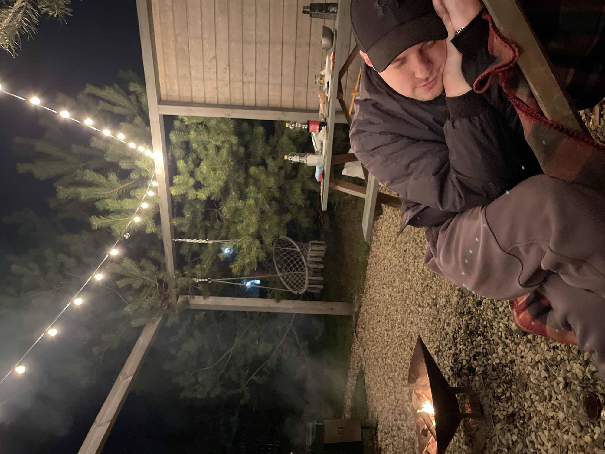 The image depicts a man resting outdoors in Литвиново, Russia. He is wearing a black baseball cap and a dark jacket, with a patterned blanket partially visible. He is seated near a fire pit, surrounded by gravel. In the background, string lights illuminate a wooden structure and greenery, including a hanging chair. A table with various items sits behind the man. The time appears to be evening.