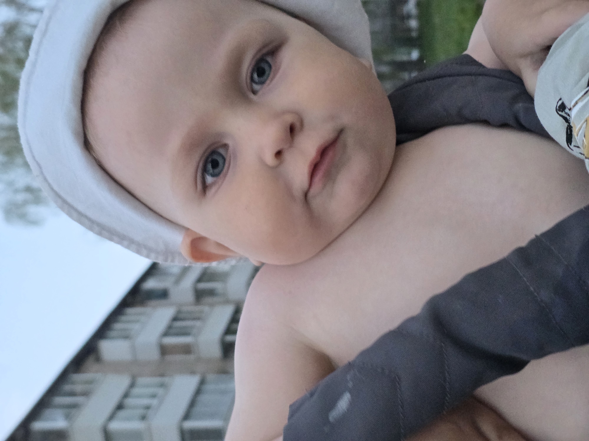 A baby with blue eyes and a white hat is being held. A building is in the background. The exact location in the city of Chaikovsky, Russia cannot be determined from this image.