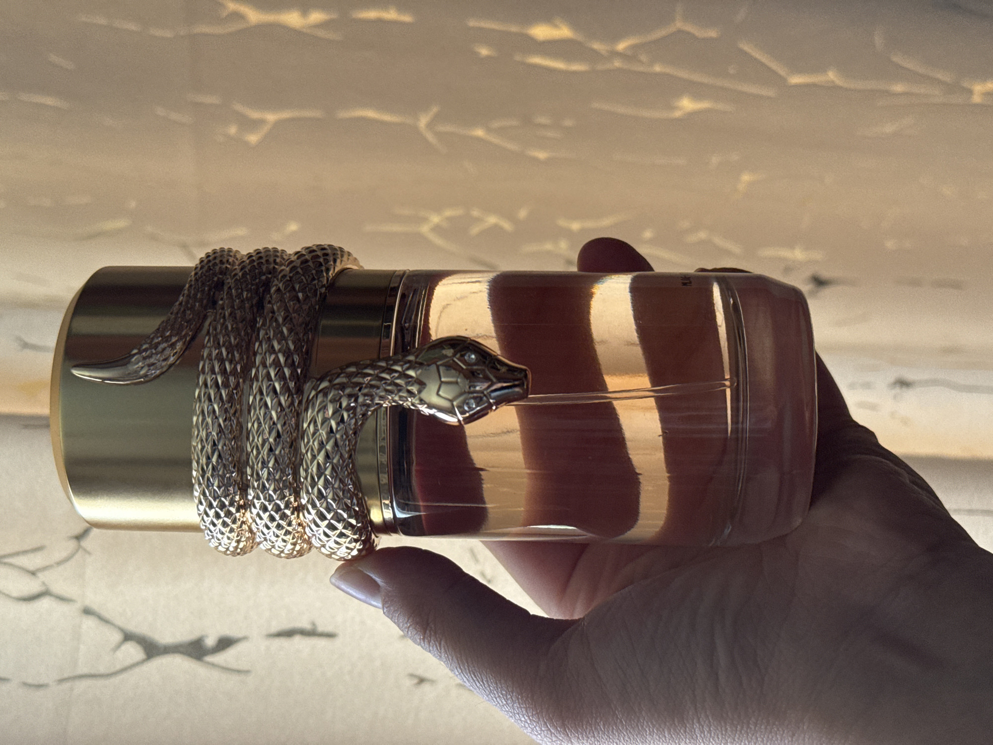 A hand holds a gold-capped perfume bottle wrapped in a decorative gold snake. The bottle contains a light orange liquid. The background consists of a sheer curtain with an abstract pattern, illuminated by light that creates dappled shadows on a wall. There is no indication of the city of Shymkent, Kazakhstan.