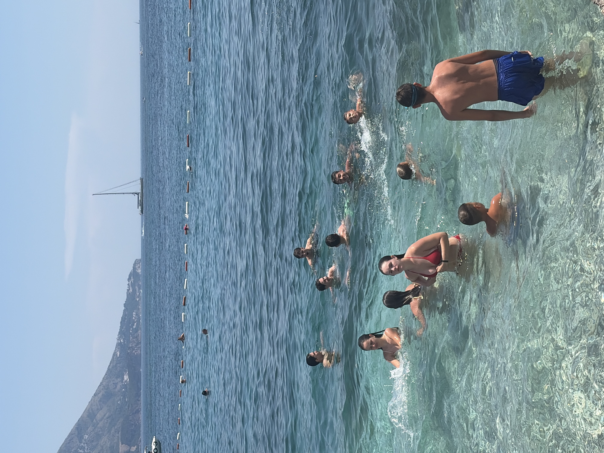 The image captures a group of people enjoying a day at the beach in Budva, Montenegro. The scene is set on a clear day with bright sunlight, reflected in the calm, turquoise waters. Several individuals, including men, women, and children, are visible swimming and wading in the sea. Some are fully submerged, while others have their upper bodies above the water, their faces expressing enjoyment and relaxation. One person in the foreground is wearing blue swim trunks and appears to be walking in the shallow water.
The water is crystal clear, allowing the rocky seabed to be seen beneath the surface. A string of buoys marks a boundary in the water, separating different areas of the beach or swimming zone. In the background, a mountainous coastline rises dramatically from the sea, characteristic of the Budva region. A sailboat or yacht can be seen in the distance, adding to the idyllic coastal scenery. The overall atmosphere is one of leisure and summer fun. No visible text is present in the image.