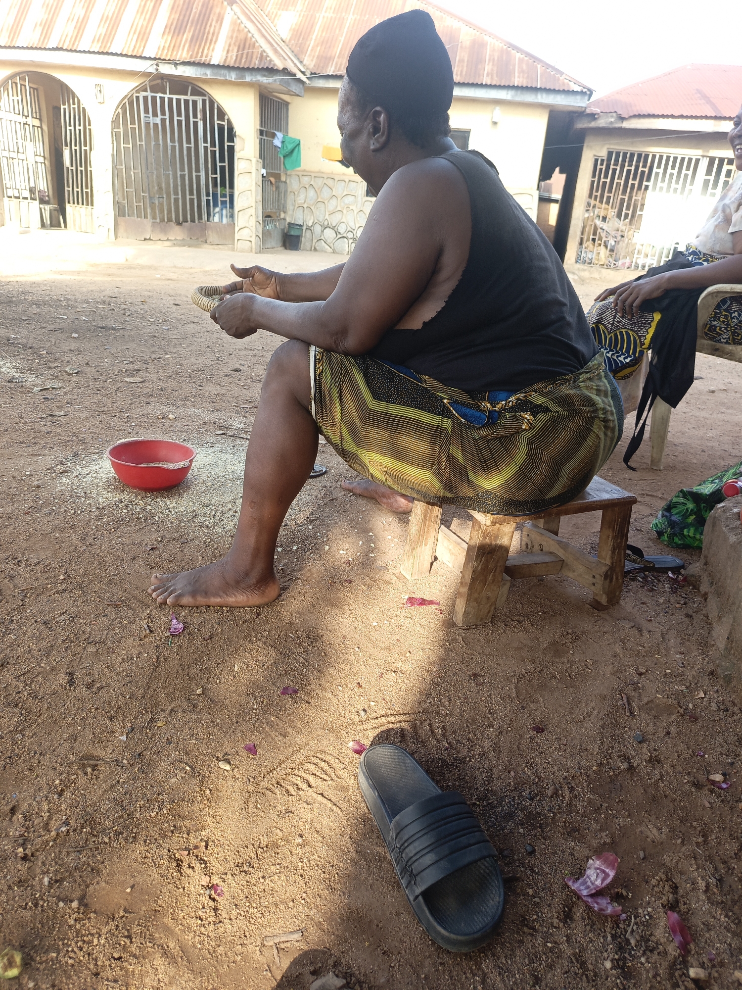 The image shows a woman sitting on a small wooden stool outdoors in Akwanga, Nigeria. She is wearing a black tank top, a patterned yellow and green wrap skirt, and a black head covering. She is holding a shallow, woven basket in her hands, likely sorting grains. A red bowl containing grains sits on the ground nearby, and scattered grains are visible around it. The ground is sandy and contains small rocks and some scattered red flower petals. A black sandal lies on the ground in the foreground. In the background, there are houses with rusty corrugated metal roofs. Another person is partially visible seated on a chair behind the woman. The scene appears to be a domestic setting with the woman engaged in food preparation.