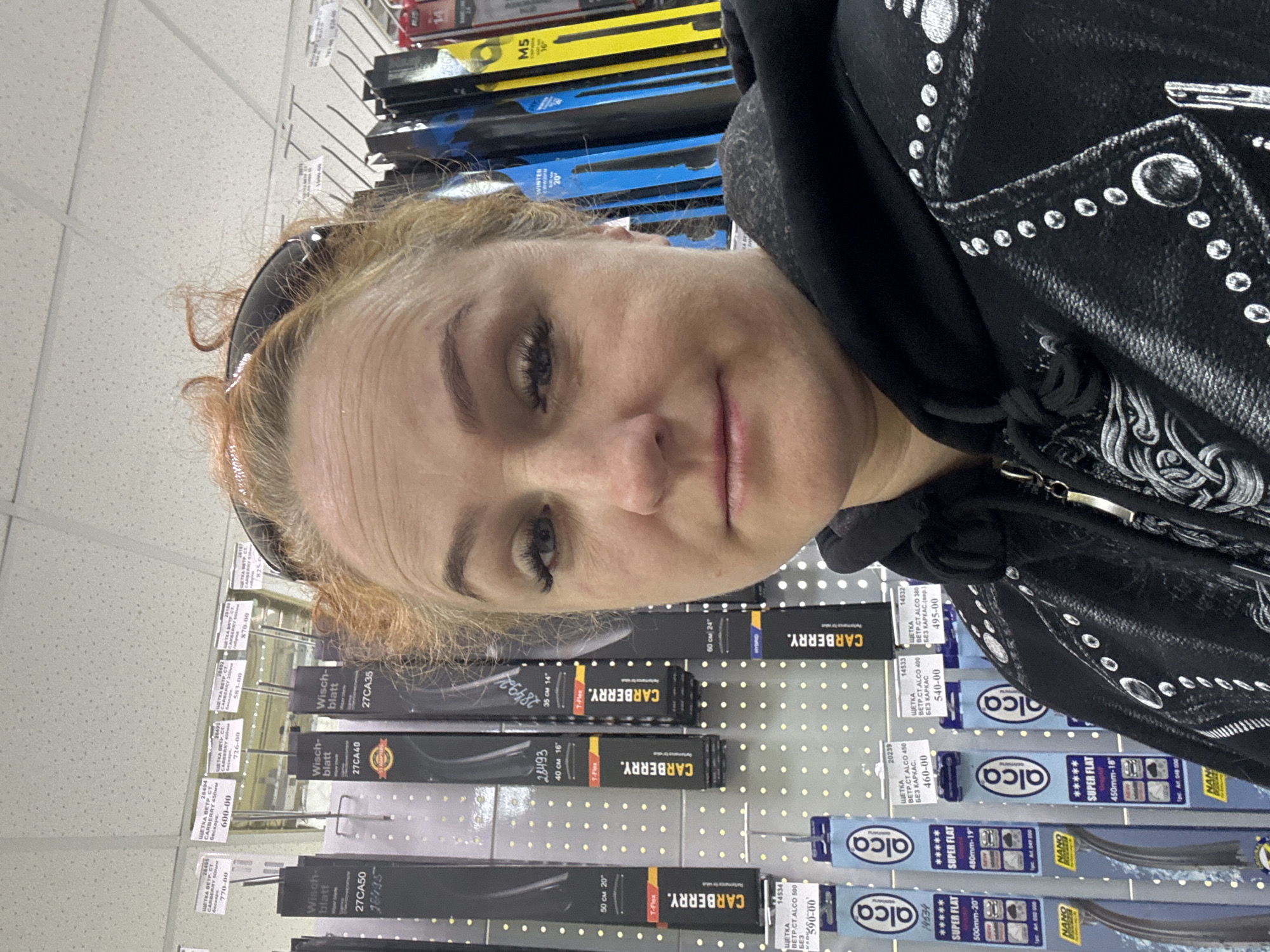 A woman is standing in front of a display of wiper blades, likely in an automotive or hardware store. The wiper blades are of various brands, including "CARBERRY" and "alca," and they are displayed on a perforated metal panel. The labels are in Russian and include the prices for the items. The store could be in the city of Chaikovsky, Russia, given the information on the labels. The woman is wearing a black jacket with silver embellishments. She has blonde hair and is looking at the camera. There is shelving above the wiper blades with additional products. The lighting is bright and the scene appears to be indoors.