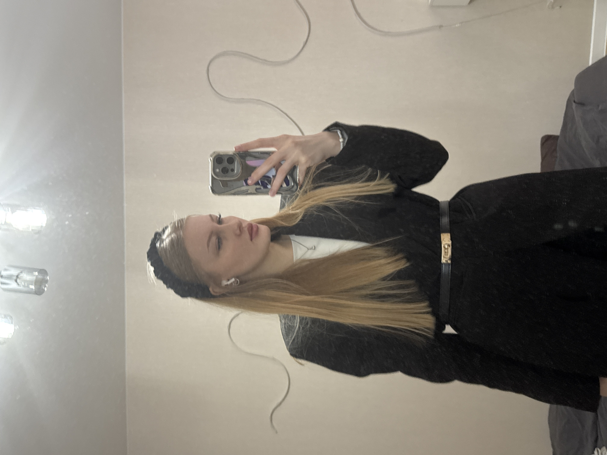 The image shows a young woman taking a selfie in what appears to be an indoor setting, possibly a room in an apartment or house. The room has white walls with electrical wiring or decorative elements visible near the ceiling. The woman is wearing a black headband, a black blazer, and a black belt with a gold buckle. She has long, blonde hair. The setting is not identifiably the city of Perm, Russia; there are no elements in the photo which establish a specific location. The woman is holding a smartphone to take the photo, and a grey bed or bedding is partially visible in the background. The lighting seems to come from overhead fixtures.