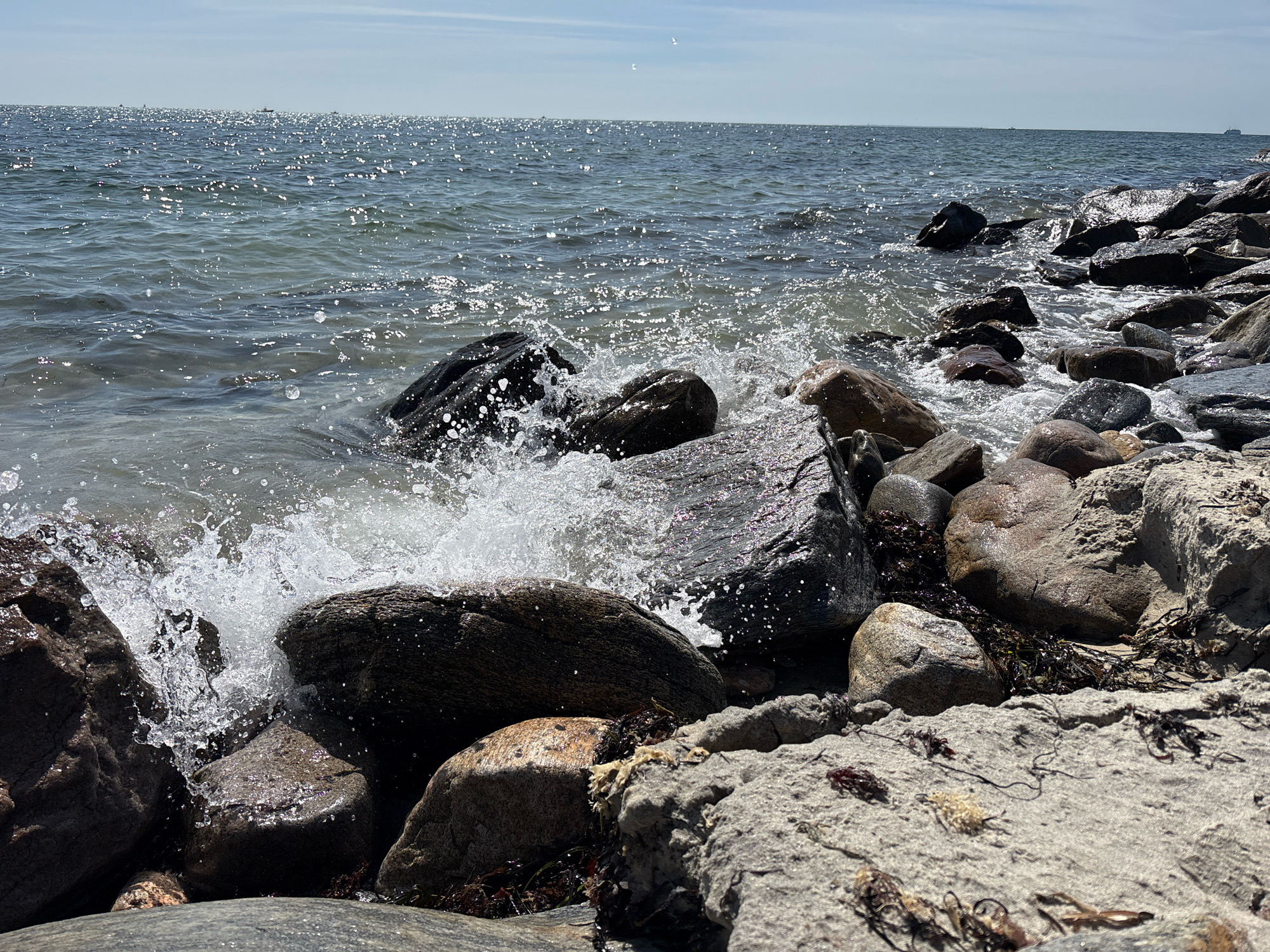The image shows a rocky shoreline with waves crashing against the rocks. The water is choppy and sparkling in the sunlight. There are many different sizes and shapes of rocks, some smooth and rounded, others jagged and rough. Some of the rocks are covered in seaweed. The sky is blue with a few white clouds. In the distance, there are a few boats on the water. This appears to be a natural, undeveloped coastline. There are no buildings, infrastructure, or signs of human habitation visible in the immediate scene, suggesting it may be a park, nature preserve, or a less developed area within Waterford, United States of America.