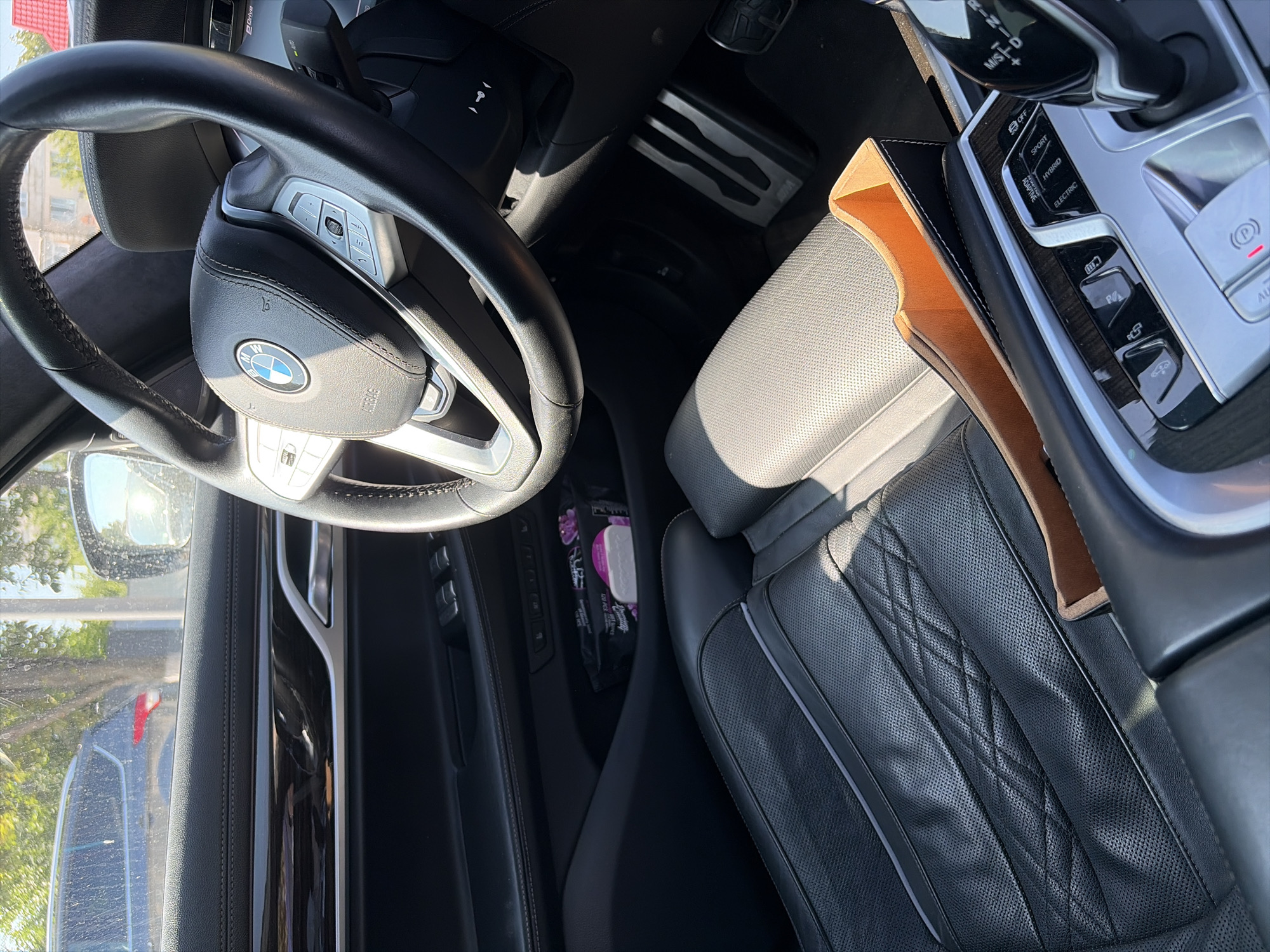 This is a close-up, interior shot of a modern car, likely taken during daylight with sunlight casting shadows. The focus is on the driver's side, featuring a black leather steering wheel with a BMW emblem, part of the dashboard with various buttons and a gear shifter, and a perforated black leather seat. A brown leather-lined organizer is placed in the center console area. In the background, through the car window, a dark-colored SUV is visible parked outdoors, along with trees and a building with a red roof, suggesting an urban or suburban setting. The image does not provide any specific indicators of the location within the city of Criuleni, Moldova. There are no people or actions depicted in the scene.