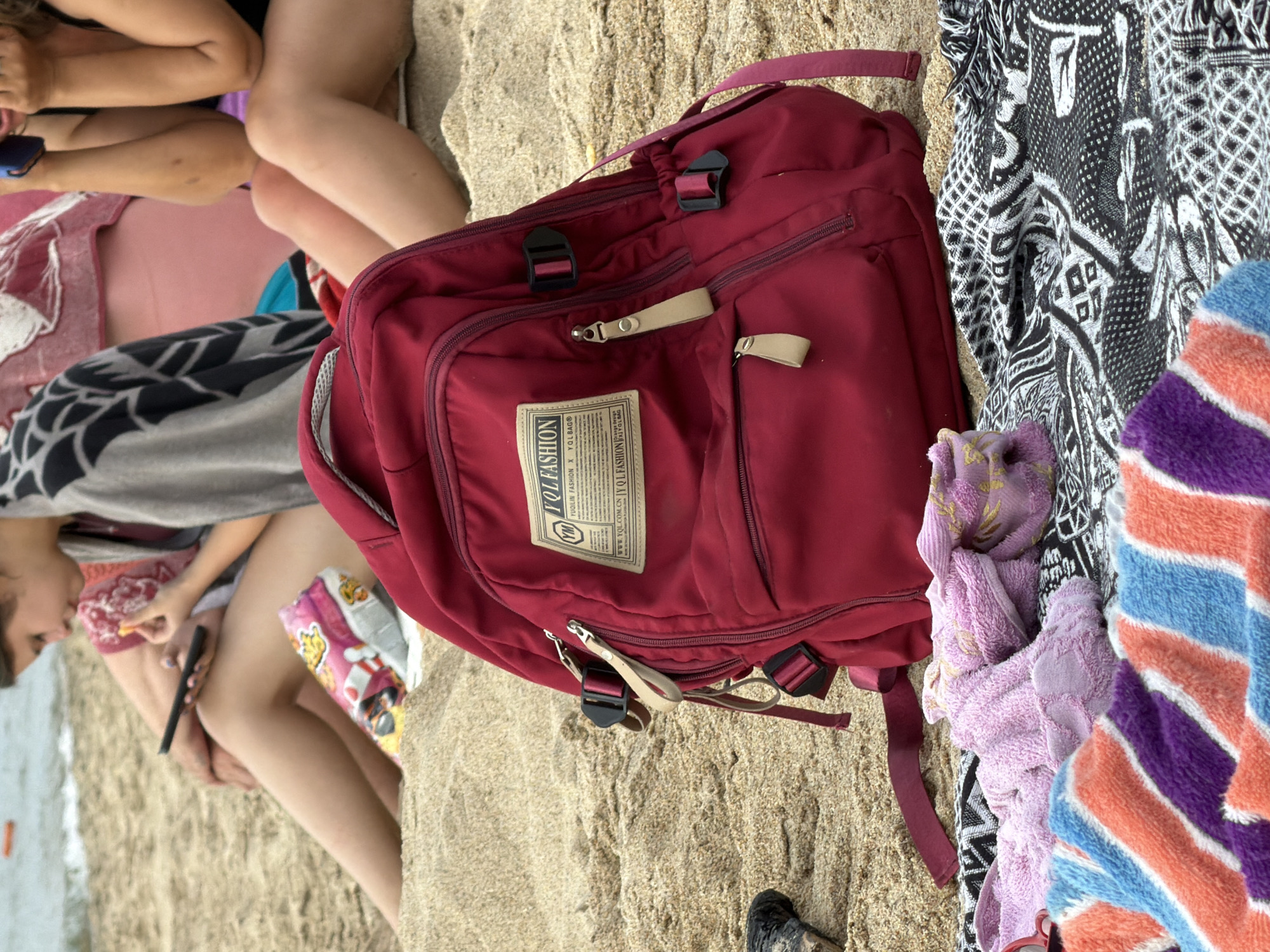 A red backpack rests on a sandy beach next to a black and white patterned blanket and a striped towel. In the background, two people are sitting on the sand. One person is looking at their phone while eating. The other person's legs are visible, and they are holding a blue object. The water is visible in the distance. This scene takes place in Goryachinsk, Russia.