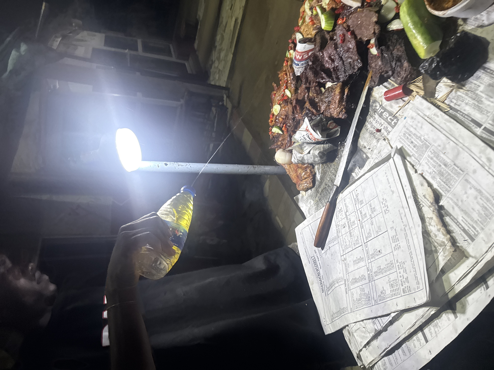 In Abuja, Nigeria, a person is pouring a yellow liquid from a clear plastic bottle onto a spread of grilled meats. The scene takes place outdoors at night, illuminated by a bright overhead light and a street lamp. A stack of newspapers, one with the masthead "NIGERIAN NEWSDIRECT," lies on the surface where the meat is displayed. The meat appears to be seasoned with spices and garnished with lime wedges and chili peppers. Several bottles and food items are also visible on the table, along with toothpicks and what looks like charcoal.