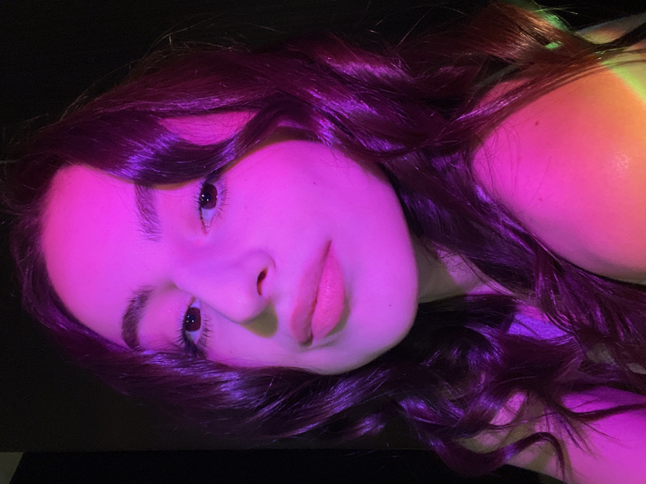 A woman's face dominates the frame, bathed in neon pink light. Her dark hair is wavy and glossy, contrasting with her fair skin. She gazes towards the left with dark eyes and softly defined eyebrows. Her lips are full and slightly parted. The background is dark and indistinct, except for a small area of greenish light near her shoulder. The overall effect is vibrant and striking.
