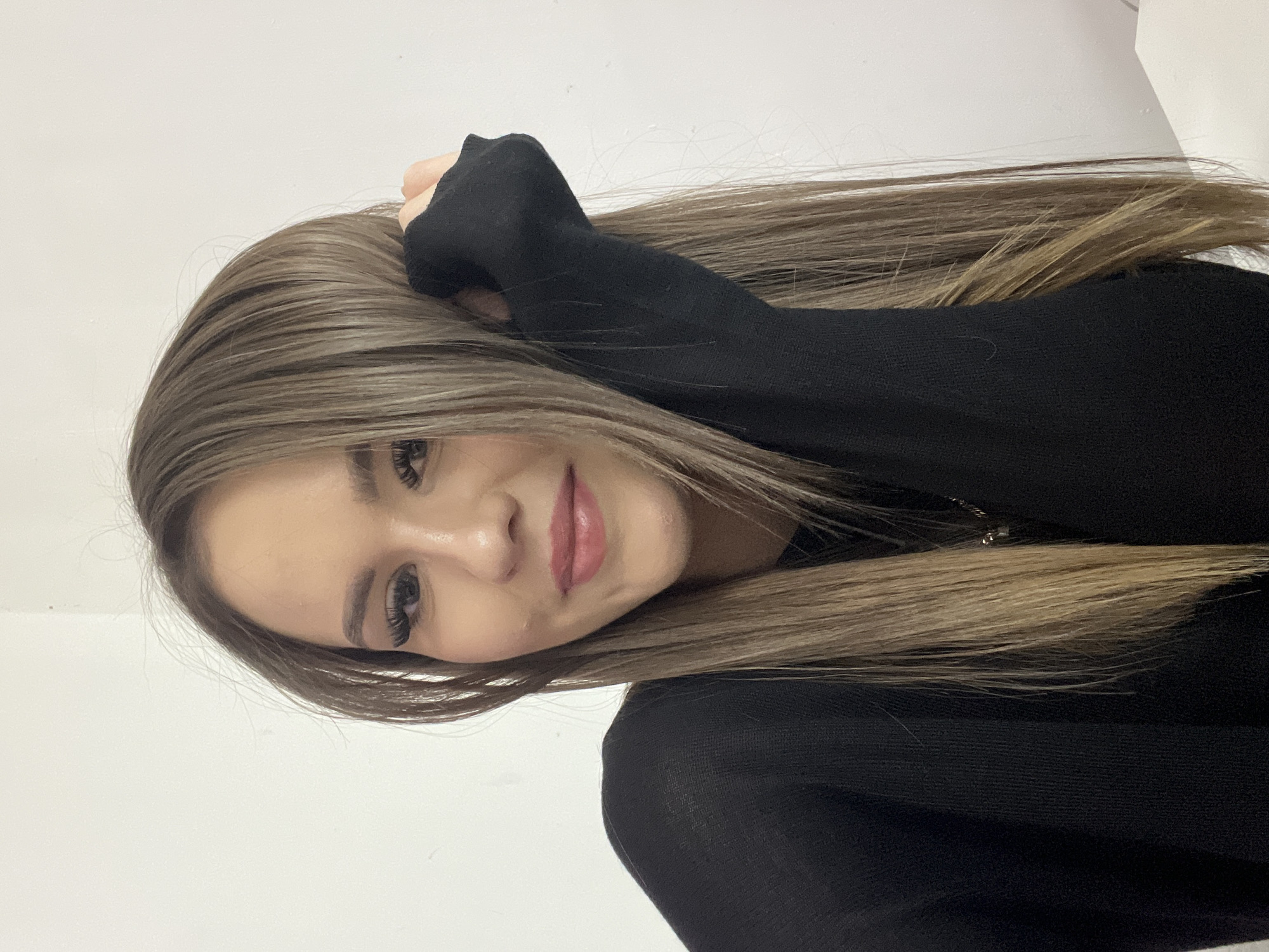 A young woman with long, straight brown hair and light-colored eyes is posing for a selfie. She is wearing a black long-sleeved shirt. Her left hand is raised and touching her hair. She is smiling softly. The background is a plain white wall. The image is cropped to focus on the woman's face and upper body. There is no identifiable information about the location in Noviy, Russia.