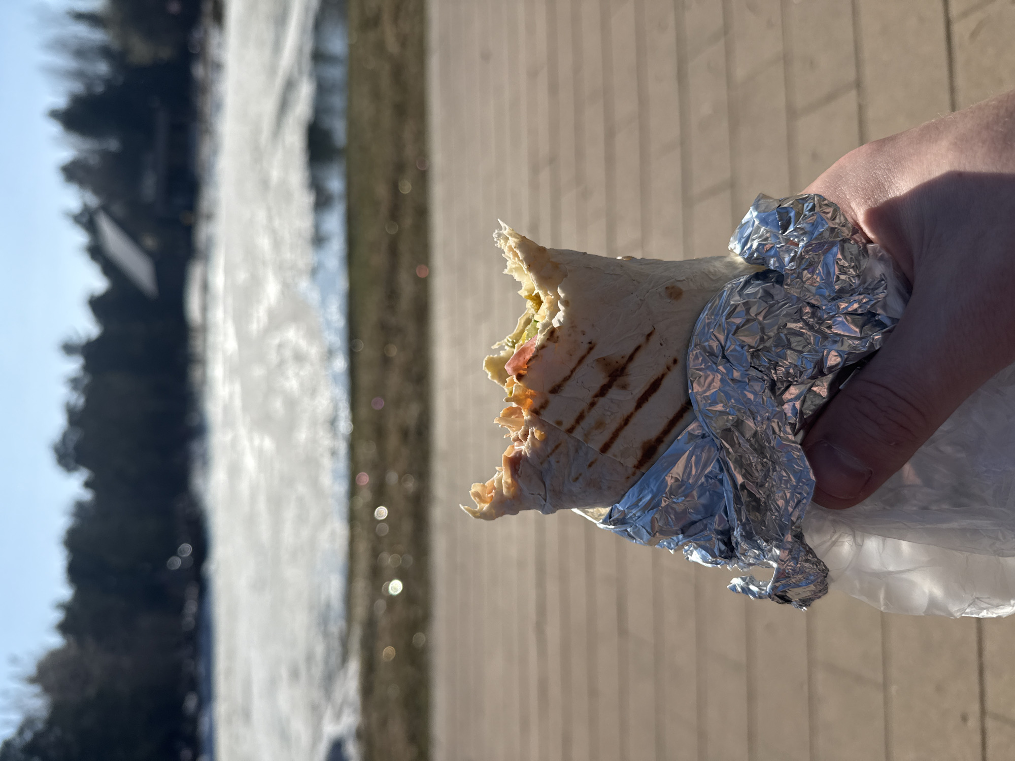 A half-eaten wrap or shawarma, partially wrapped in foil, is held in a person's hand against an outdoor background. The location appears to be Moscow, Russia, judging by the style of the urban park with a waterfall feature and light-colored wooden boardwalk. The food item reveals a portion of its filling, hinting at ingredients like vegetables and meat. The scene is well-lit, suggesting it is daytime. The perspective is from a point-of-view shot as though the photographer is about to eat the wrap.