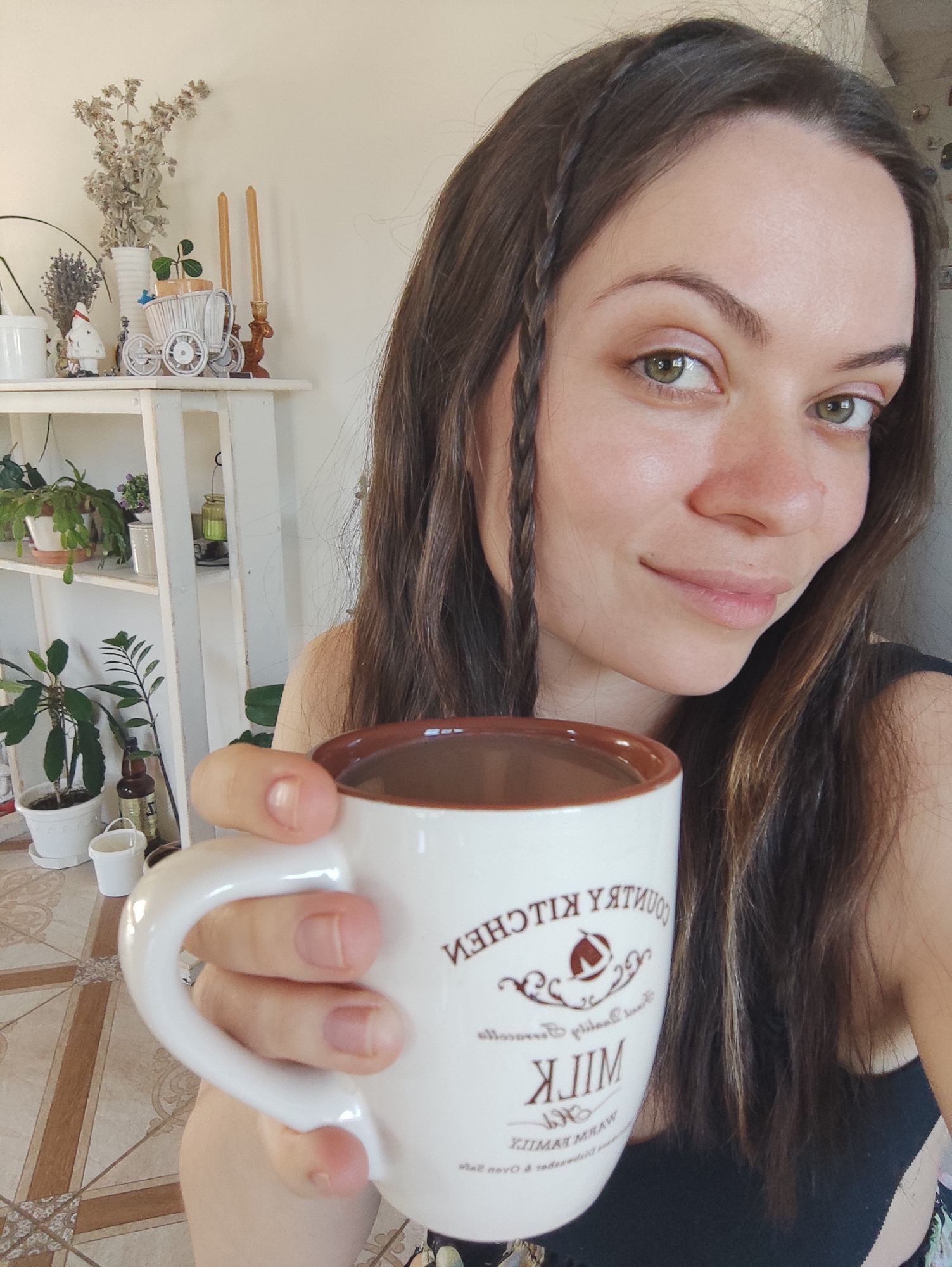 A brightly lit, vertical shot captures a woman holding a white mug, looking directly at the camera with a gentle, soft smile. Her long, dark brown hair cascades over her shoulders, with thin braids framing her face. She has striking green eyes and a natural complexion.
The white ceramic mug, held by her right hand, features a brown rim and visible text in a rustic font, which is mirrored in the image. The text reads "COUNTRY KITCHEN" at the top, an emblem resembling a house in the middle, and "MILK" below it, followed by smaller, less legible text including "WARM FAMILY". The mug appears to contain a dark beverage, possibly coffee or hot chocolate.
In the background, a white multi-tiered shelf unit serves as a display for various decorative items and plants. On the top shelf, a dried floral arrangement in a white vase stands next to two tall, slender candles in a decorative holder. A small white ornamental wagon and a figurine, possibly a small gnome or Santa, are also visible. The lower shelves are adorned with several potted green plants, a brown bottle, and a small white bucket. The floor in the foreground features a brown and beige patterned tile design.
The scene takes place indoors, likely during the daytime, given the soft, natural illumination. The overall mood conveyed is one of calm and contentment. This indoor setting is situated in Фоки, Russia.