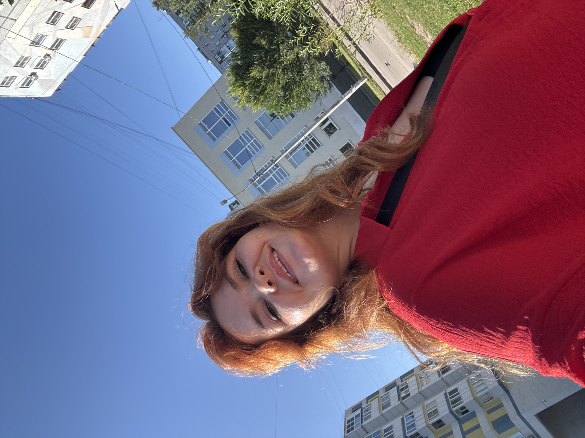 An outdoor, low-angle, full-body, selfie-style photograph features a young woman with reddish-brown, wavy hair and fair skin. She is smiling and looking at the camera, with her face illuminated by sunlight. She is wearing a bright red, oversized garment with a black band visible across her chest. The background consists of a clear, bright blue sky with several power lines crisscrossing it. In the middle ground, two multi-story apartment buildings are visible, angled against the sky. One building on the left has a colorful facade with yellow and blue accents and many small windows. The building on the right is lighter in color, possibly beige or light grey, with a geometric pattern and numerous windows. Trees and a sidewalk are also present. The overall impression is a sunny day in a residential urban area, possibly Nizhny Novgorod, Russia.