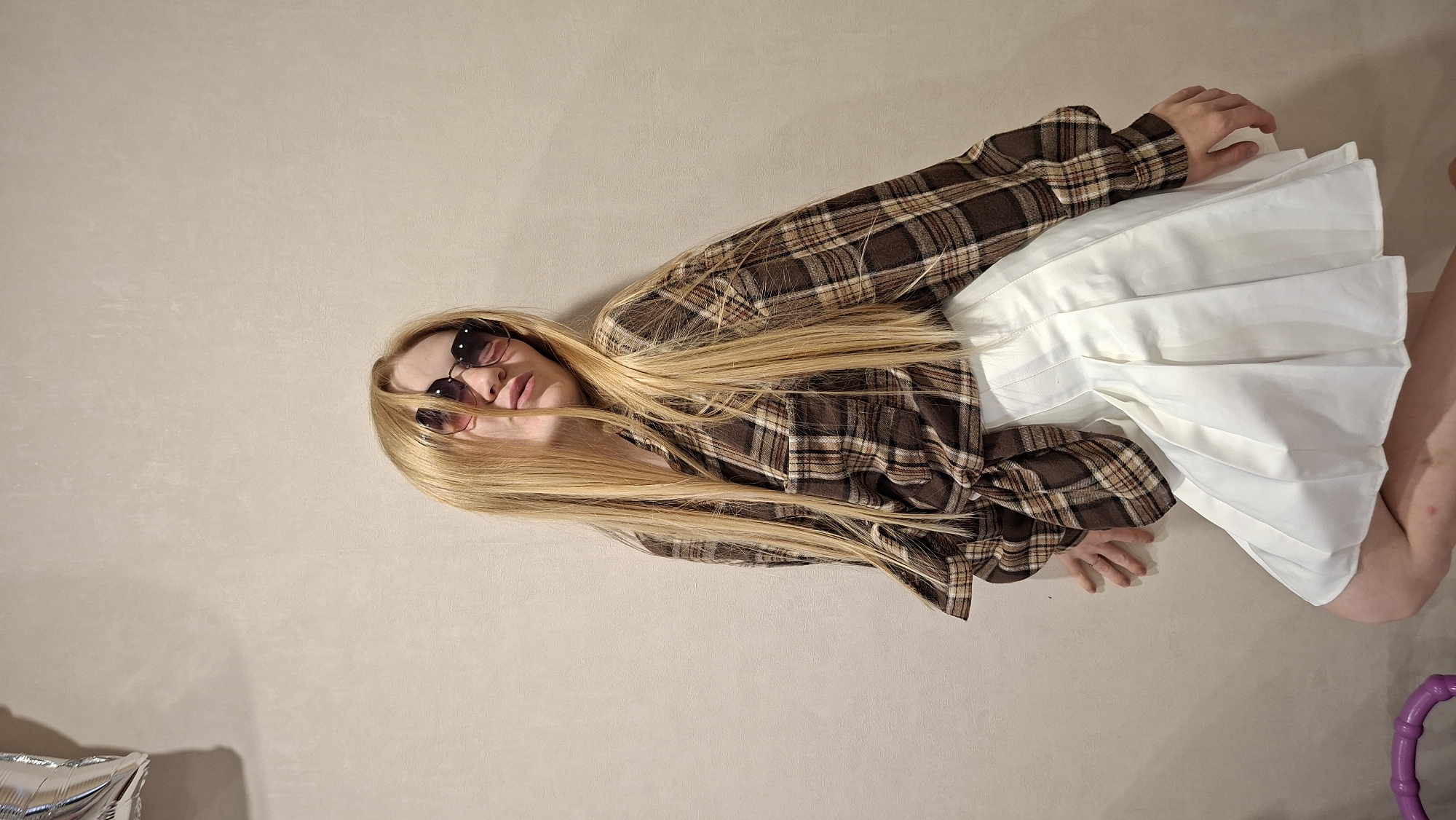 A young blonde woman with long hair, wearing sunglasses, a brown plaid shirt, and a white pleated skirt, is lying down on a light-colored floor. Her body is positioned diagonally across the frame, with her head towards the upper right corner. Her right hand is resting on her skirt. Her facial expression is relaxed. The scene is indoors, with a plain, textured wall as the background. To the lower left, a portion of a silver balloon is visible, and to the lower right, a glimpse of a purple object can be seen. The lighting suggests it is daytime, possibly indoors with artificial light or natural light coming from a window. The overall mood is casual and relaxed. No visible text is present.