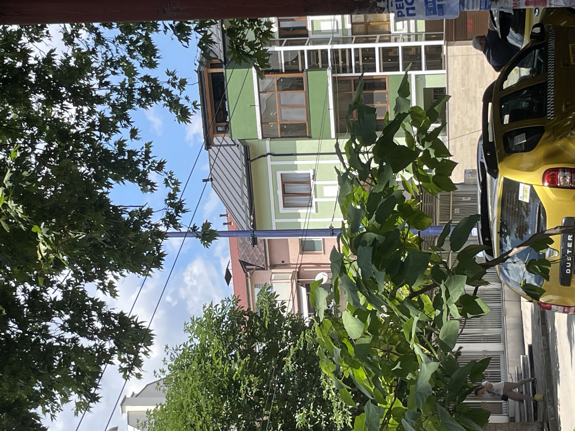 This is a vertical, eye-level shot of an urban street scene, partially obscured by foliage. A vibrant green building with multiple stories and white trim is prominent. It features several large windows, some with wooden frames and what appear to be reflective coverings. Above the green building, a section of a lighter-colored building is visible, with a dark-pitched roof. Further back, a more modern building with a grid-like facade of white and glass elements rises.
In the foreground, lush green trees with broad leaves dominate the left and right sides of the frame, casting shadows and adding a natural element to the man-made environment. A bright yellow vehicle, possibly a taxi due to the checkered pattern on its side and "DUSTER" text on the rear, is parked on the right side of the street, facing away from the camera. A person, partially visible, is standing near the vehicle. Another person is seen walking on the sidewalk in front of a building on the right.
The sky above is a clear blue with scattered white clouds, suggesting fair weather. Several power or telephone lines crisscross the upper portion of the image. The overall impression is of a sunny day in a built-up area, where nature and urban architecture coexist. The orientation of the photo is rotated 90 degrees counter-clockwise from standard.
