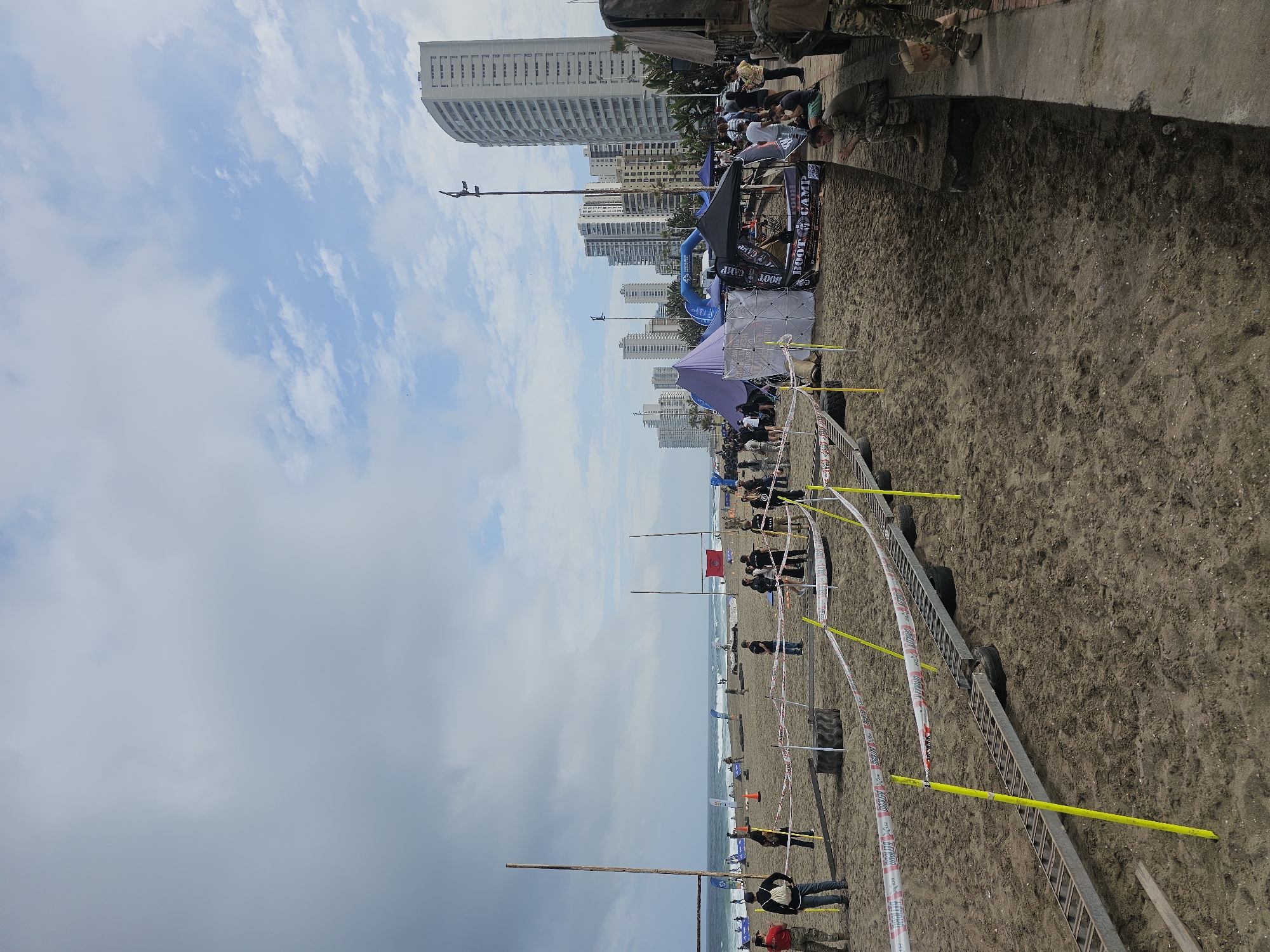 The image depicts a beach scene in Iquique, Chile, likely during an outdoor event or competition. A sandy beach stretches towards the ocean under a cloudy sky. In the foreground, there's an obstacle course set up on the sand, featuring a metal ramp-like structure and various barriers. Yellow poles and red and white tape demarcate the course. Several individuals, some in athletic or military-style clothing, are present, either participating in or observing the event. Tents and structures are visible in the middle ground, one of which appears to be a "BOOT CAMP" registration area. In the background, the city skyline of Iquique is dominated by tall residential buildings and palm trees. The ocean with moderate waves is visible on the left. The overall atmosphere suggests an organized outdoor activity on a potentially overcast day.
