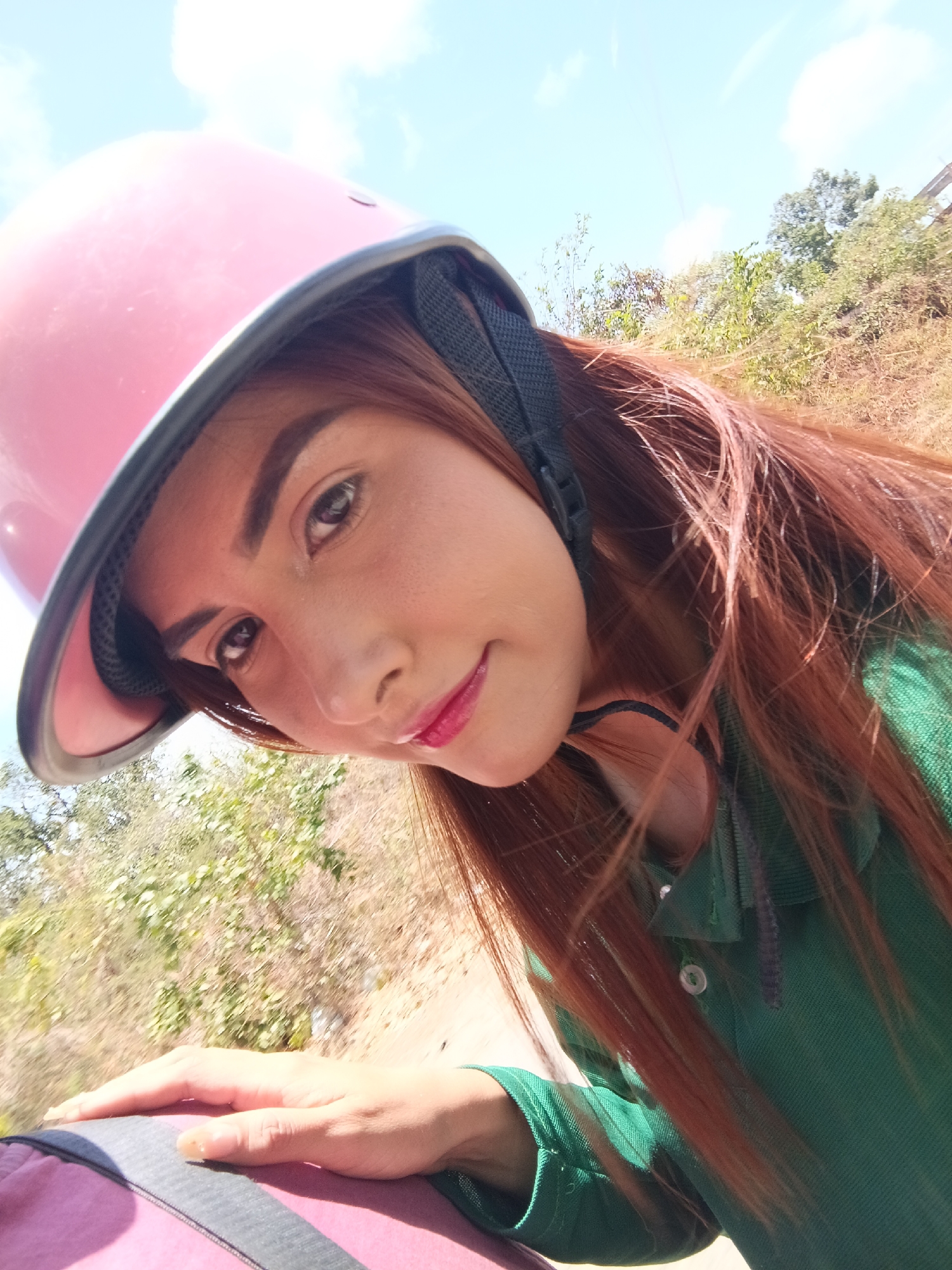 A woman is taking a selfie outdoors, likely while riding on a motorcycle or similar vehicle. She is wearing a pink helmet with a black strap and a green polo shirt. Her long, reddish-brown hair is visible, partially tucked under the helmet and partially blowing in the wind. She is fair-skinned with dark eyebrows and is wearing lipstick. Her left hand is visible, resting on what appears to be part of the vehicle or a bag. The background is blurry and consists of vegetation, suggesting a roadside environment. The sky is visible in the upper portion of the frame with patchy clouds. The exact location within Juan Griego, Venezuela, is not discernible from the image.