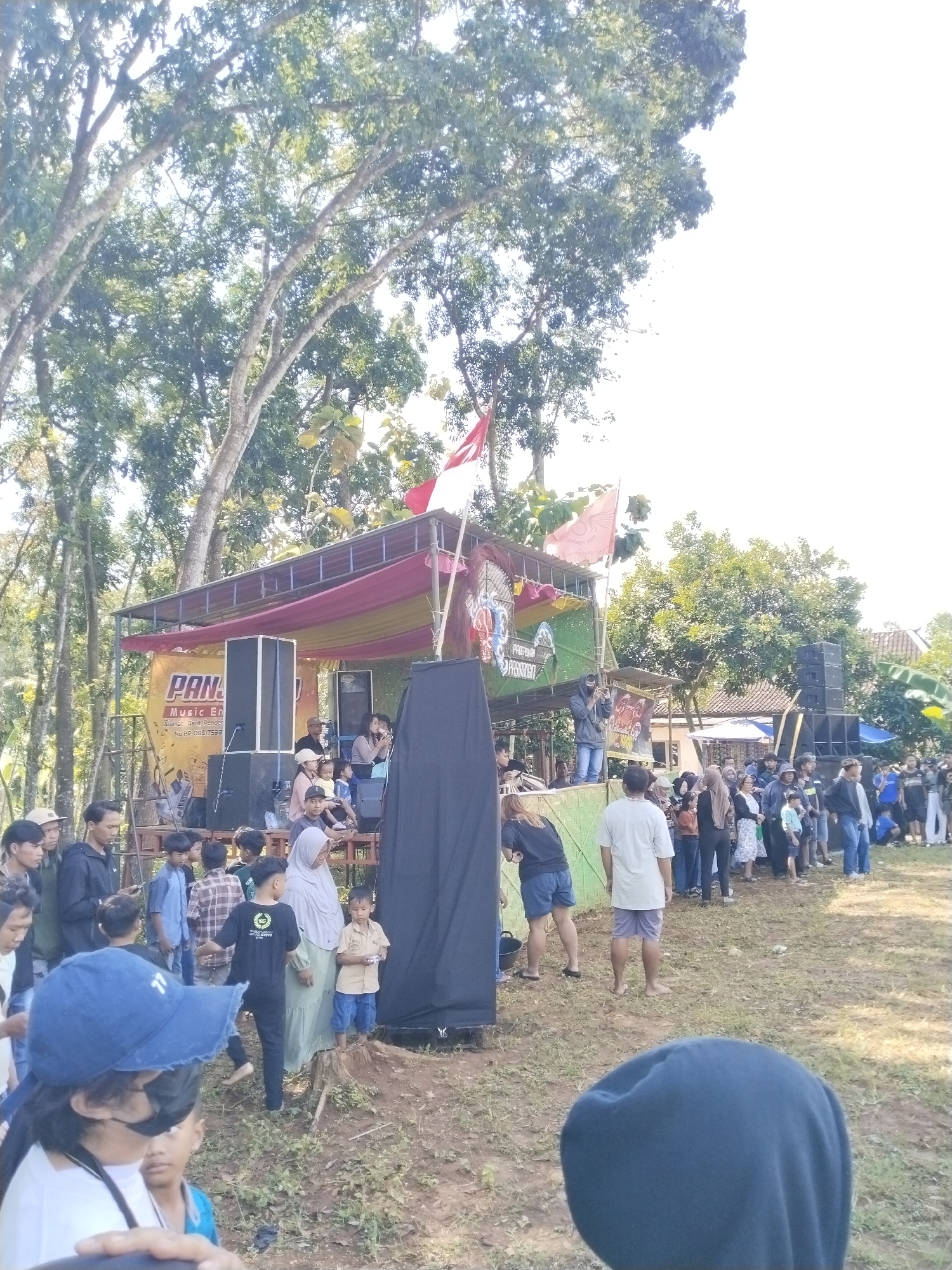 The image depicts an outdoor event, potentially a festival or community gathering, located in an unspecified city in an unknown region. A makeshift stage with a yellow and green backdrop, partially obscured by a black drape, is visible. People of various ages and attire are present, suggesting a diverse crowd. Some are standing in a line, while others are gathered around the stage. Indonesian flags are displayed atop the stage, indicating the event is likely in Indonesia. The surrounding environment consists of trees and grass, indicating a park or outdoor setting.