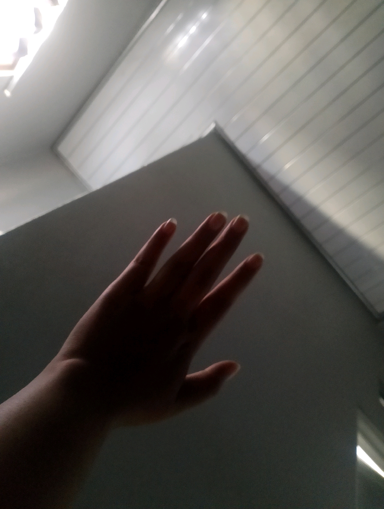 In a dimly lit indoor environment, likely a room, a left hand is positioned centrally in the frame, palm facing away from the viewer and fingers slightly spread. The hand is silhouetted against a lighter background, with subtle lighting illuminating the fingertips and the back of the hand. Above the hand, a white, corrugated ceiling is visible, with angled lines suggesting a sloped surface or a perspective view of a ceiling structure. The overall lighting is low, creating a contrast between the shadowed hand and the illuminated ceiling. No specific location within the city of Magelang, Indonesia, can be determined from this image.