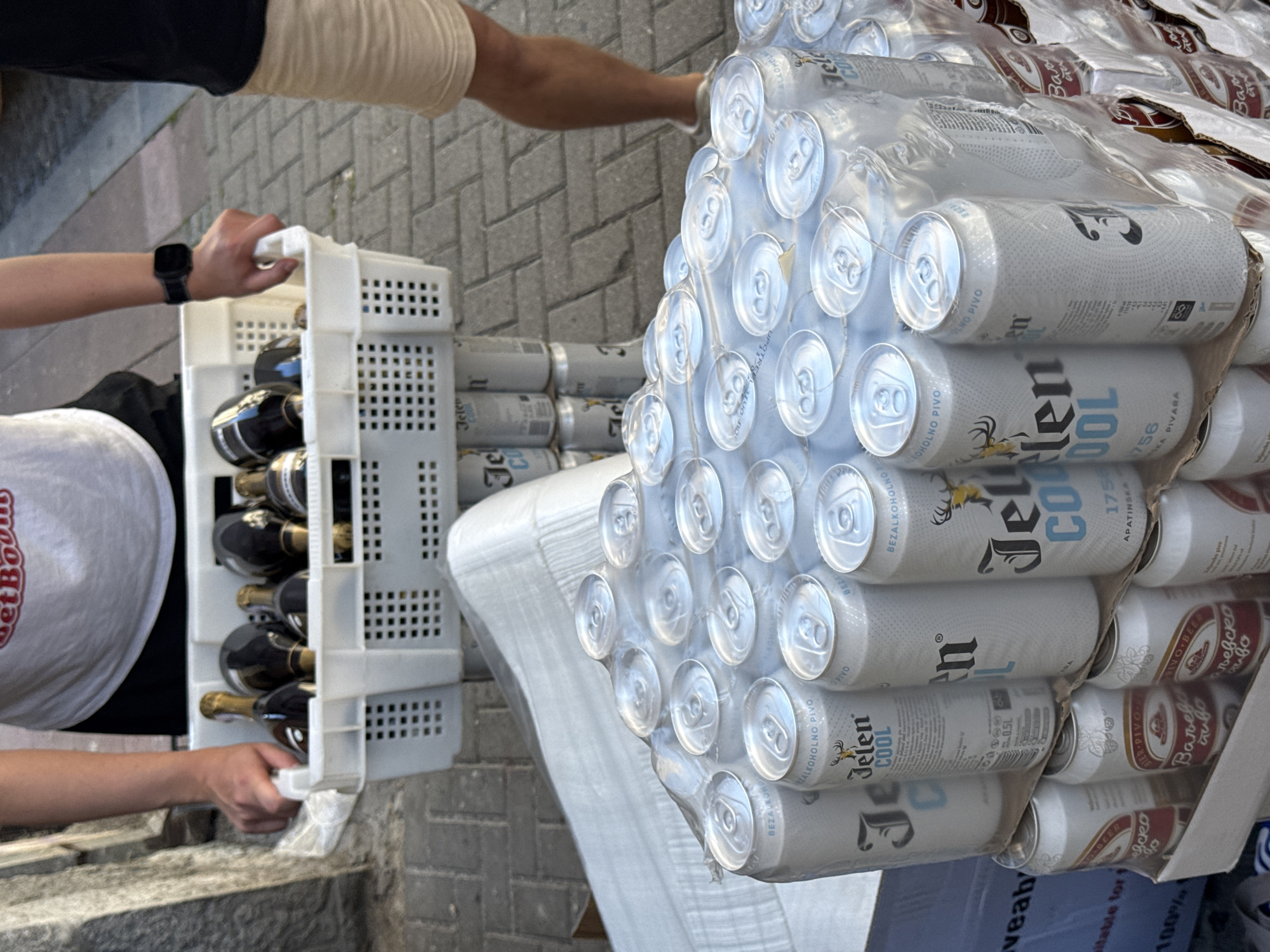 The image shows two people outside in Belgrade, Serbia, carrying crates of beverages. The person on the left is holding a white crate containing wine bottles, while the person on the right is carrying a large stack of Jelen Cool beer cans. The setting appears to be a street with brick paving.
