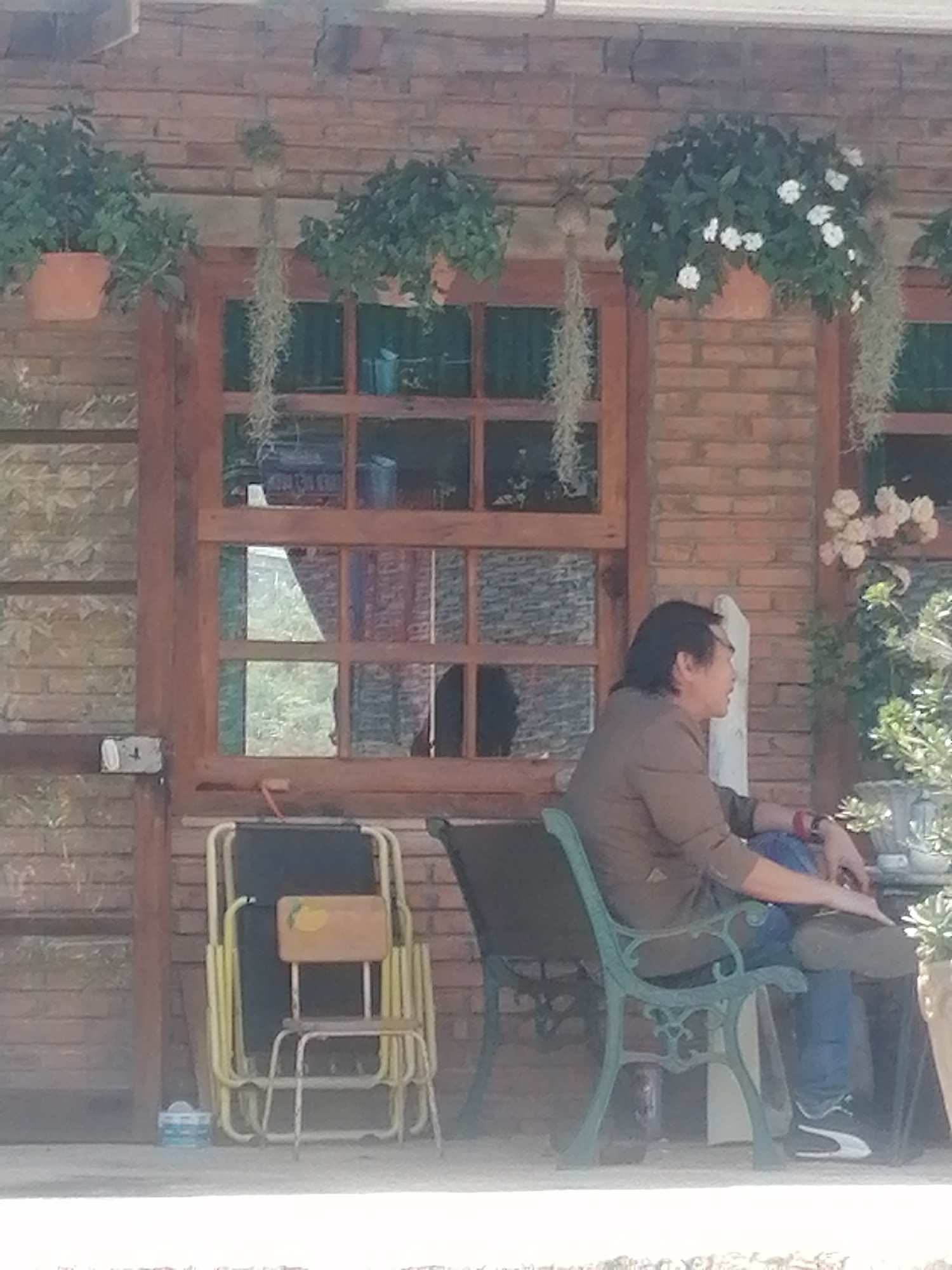 The image shows a man sitting on a green bench in front of a brick wall with a window. He is wearing a brown shirt and blue jeans, and he has long, dark hair. The window has wooden frames and is divided into panes. There are plants hanging in pots above the window, and a bouquet of flowers on the right side of the window. In front of the window, there is a stack of yellow chairs and a dark-colored object. The man's legs are crossed, and he is looking to the right. The scene appears to be outdoors, possibly on a patio or porch. The building is made of brick and has a roof over the area where the man is sitting. The overall impression is one of a relaxed, outdoor setting.