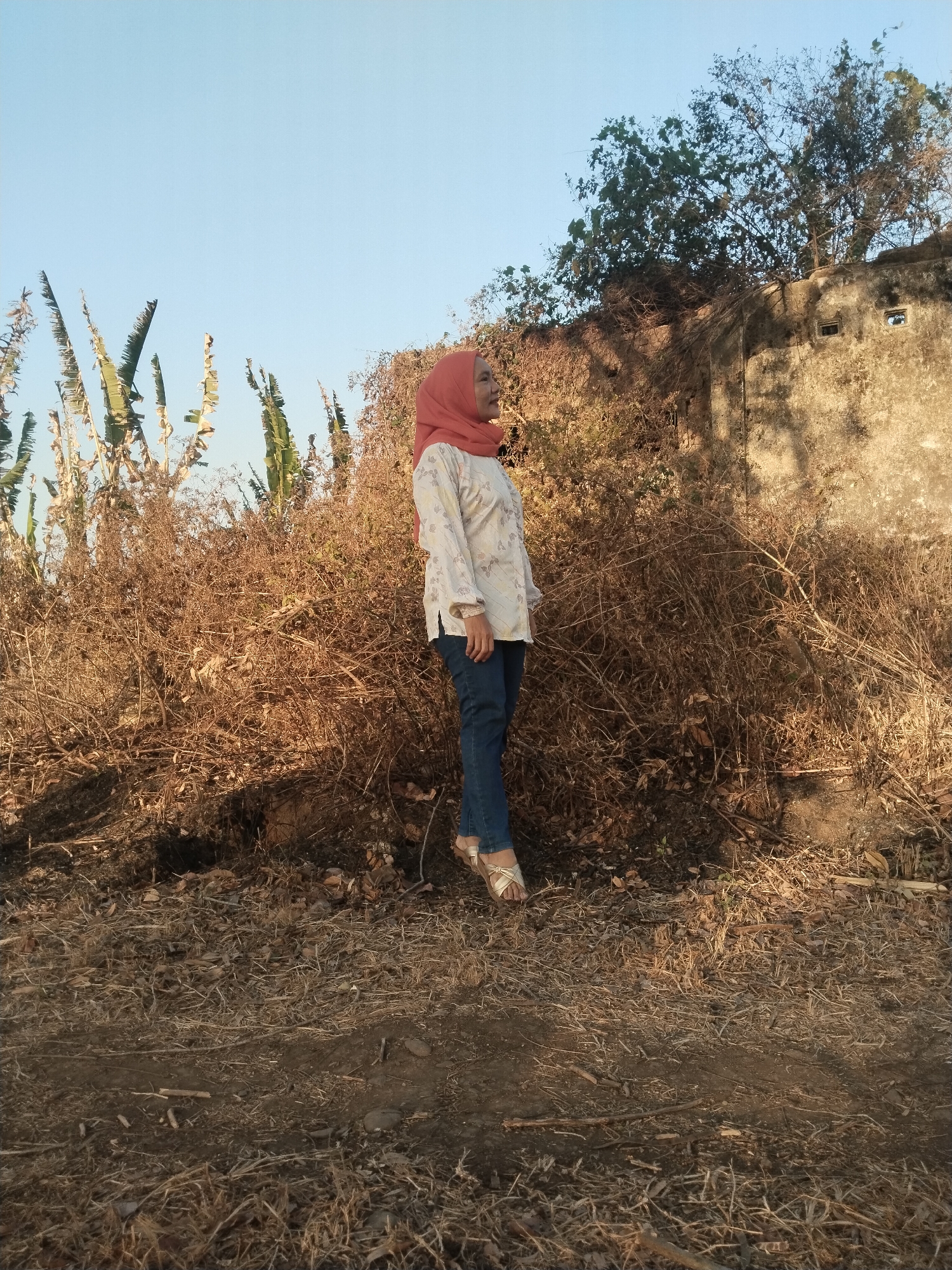 A young woman wearing a peach hijab, a white patterned shirt, and blue jeans stands facing left in a dry, overgrown area. She is barefoot and wearing sandals. Behind her, a dilapidated concrete wall with two small openings is visible, partially covered by dead foliage and trees. The ground is covered in dry grass and debris. The scene appears to be outdoors, likely in a rural or neglected urban environment.