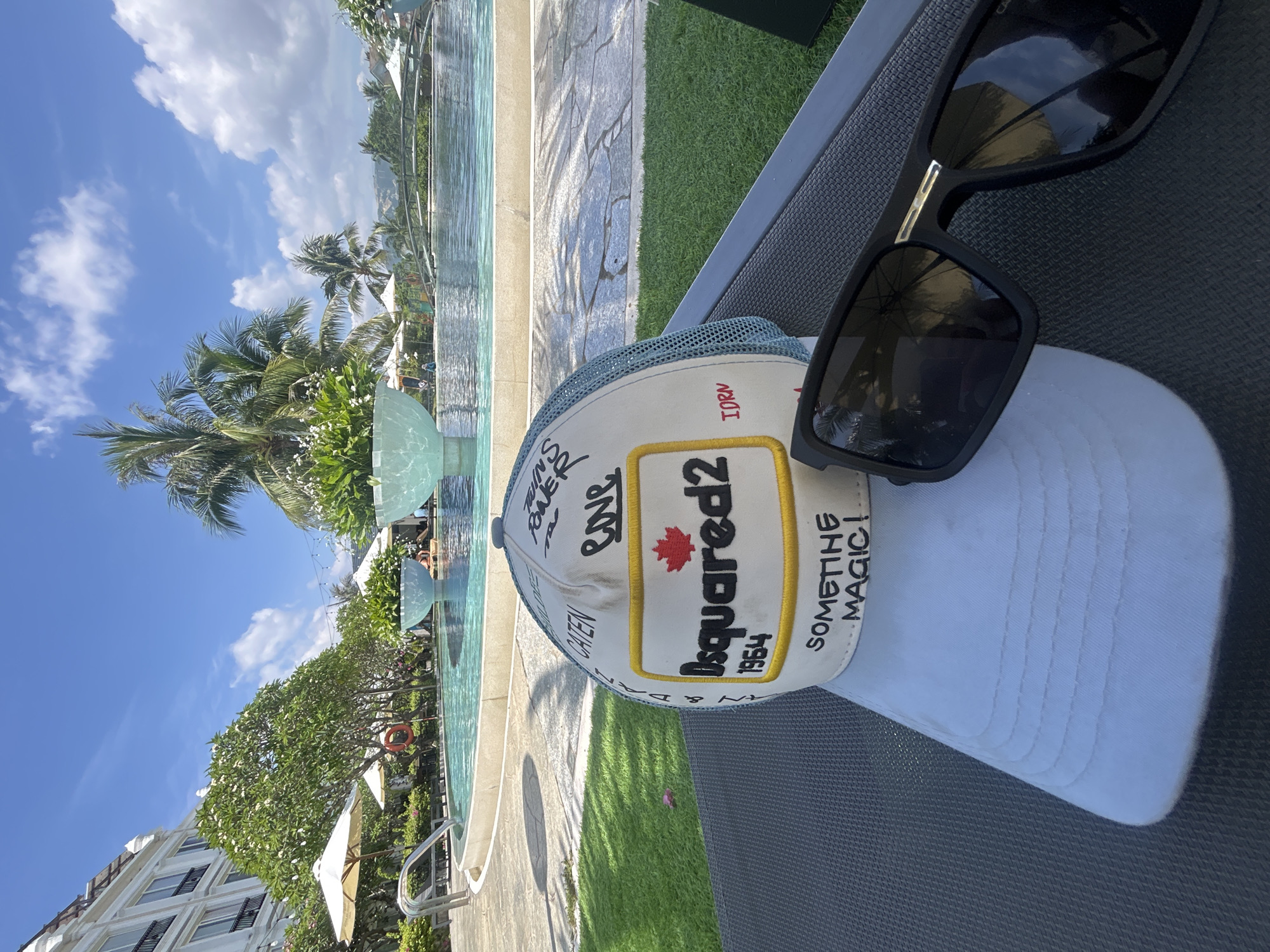 The image shows a still life composition of personal items placed on a dark-gray outdoor lounge chair with a view of an outdoor swimming pool and lush tropical vegetation. A white and light-blue Dsquared2 cap with graffiti-style writing is positioned centrally. On top of the cap rests a pair of black-framed sunglasses. The pool area is lined with green grass and palm trees, with a partial view of a white multi-story building in the background. The scene is situated in Bac Nha Trang Ward, Vietnam.