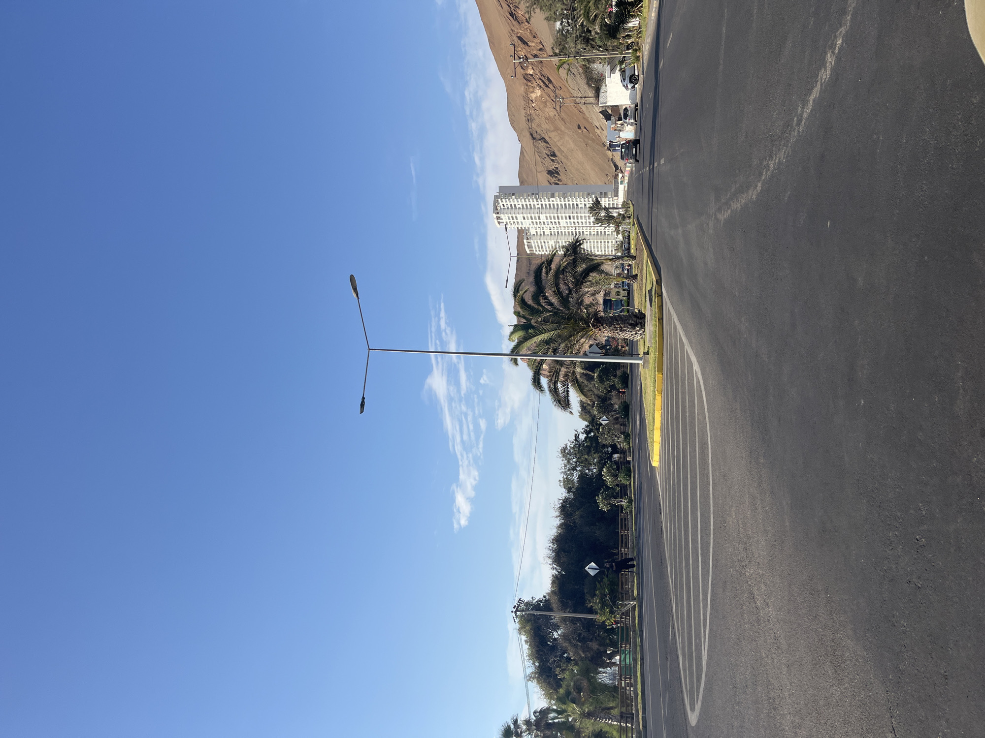 The image shows a street scene in Arica, Chile, under a clear blue sky with some wispy clouds. The street is asphalt, with white lines marking a designated area, possibly for parking or recreation. The street has a yellow curb on the left.
A tall, white building is visible in the background, positioned at the base of a brown, rocky hill or mountain. Palm trees are along the street. A light pole extends upwards from the left side of the road.
Traffic and buildings can be seen along the top of the frame. The image lacks people and detailed interactions or actions, focusing instead on the urban and natural landscape. The overall setting suggests a coastal city with mountainous terrain.