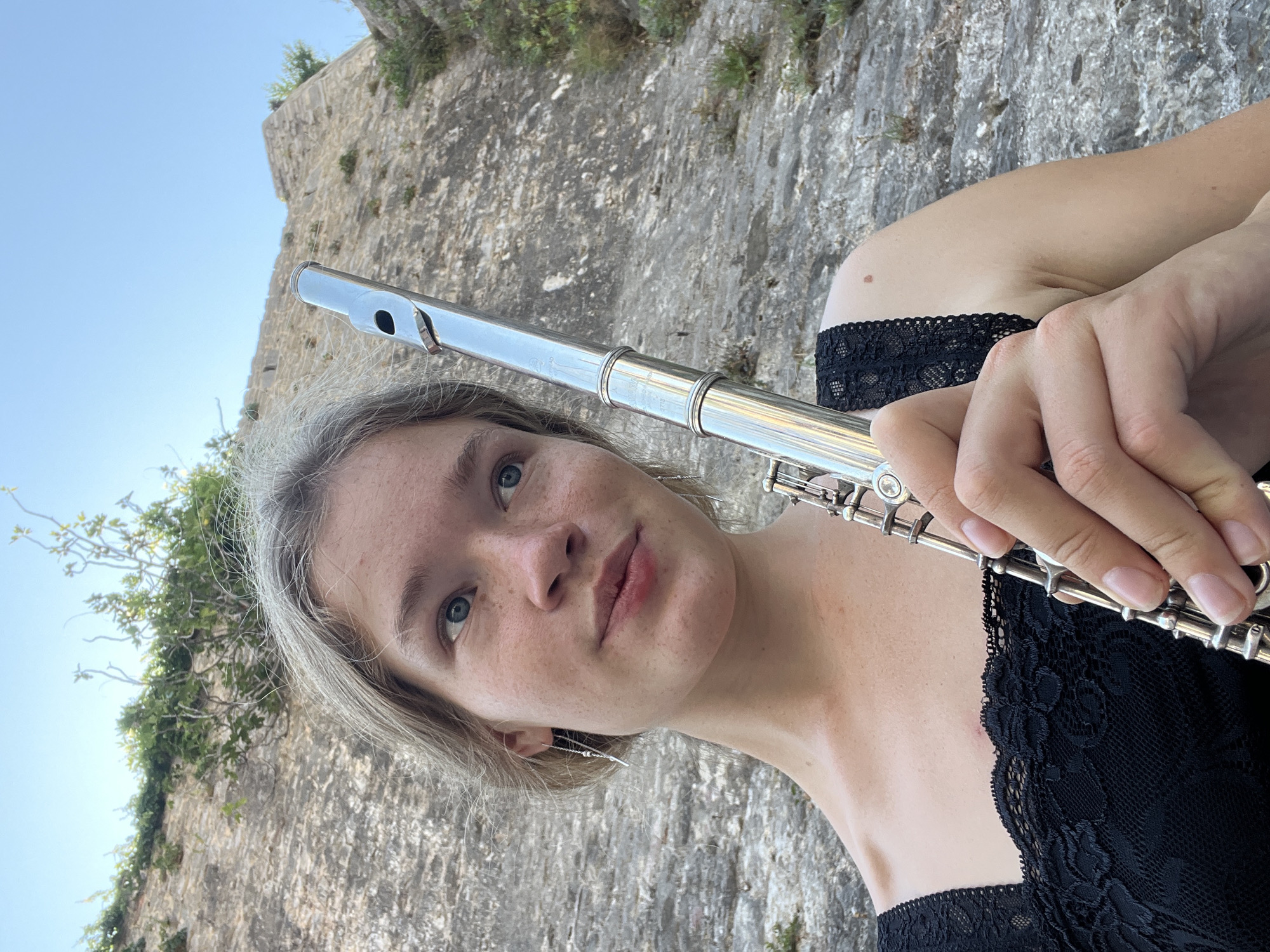 A young woman with blonde hair and blue eyes holds a silver flute. She is wearing a black lace top. The background is a rocky, sun-drenched hillside with some greenery. The scene appears to be outdoors, possibly in Budva, Montenegro, given the architecture and vegetation. She is looking slightly to her right, with a neutral expression.