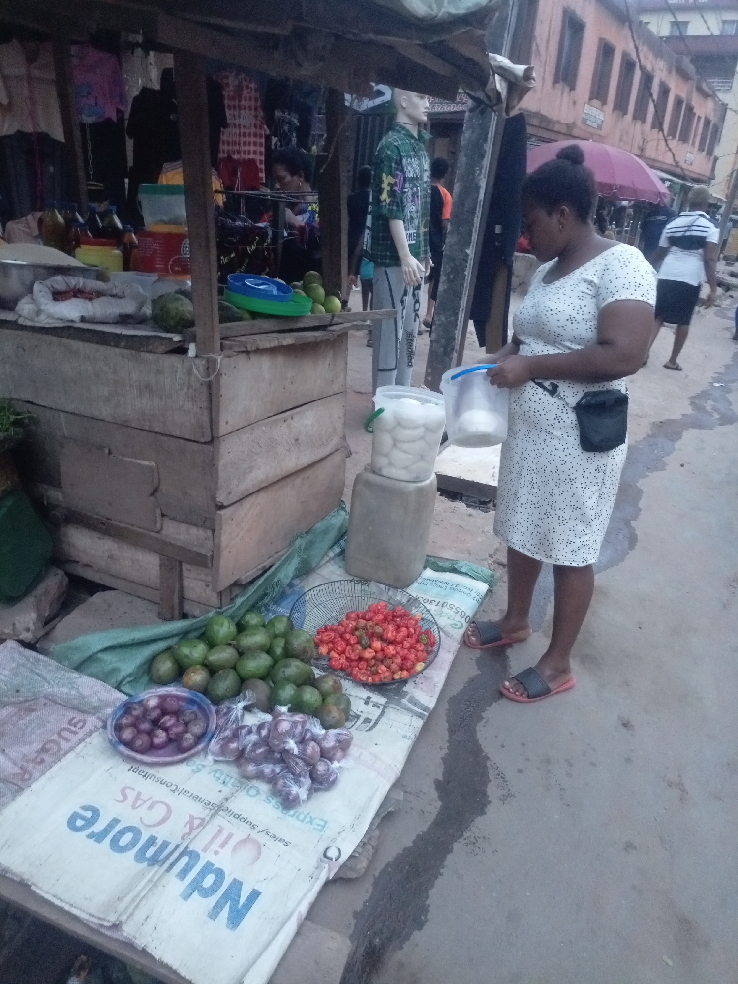 The image depicts a bustling street market scene in Onitsha, Nigeria, under what appears to be an overcast sky or late afternoon light. In the foreground, a woman in a white, short-sleeved polka-dotted dress and black slide sandals stands to the right, holding two white plastic containers or buckets. A black fanny pack is secured around her waist.
To her left, a market stall, constructed from rough wooden planks, displays various goods. On the ground in front of the stall, laid out on a large white sack, are piles of green fruits, likely avocados, a metal sieve filled with bright red chili peppers (resembling scotch bonnets), a blue bowl containing red onions, and several small plastic bags also filled with onions. The sack features prominent text including "Ndumore Oil & GAS," "Sales / Supplies General Consultant," "Onitsha Enugu Exp," and a phone number "065501303." The word "SUGAR" is also partially visible.
On the wooden stall itself, several clear bottles containing amber liquid (possibly palm oil) are visible, along with various plastic bowls and containers, one holding white, dough-like items. A mannequin, dressed in a green patterned shirt and grey pants with "Stediea" partially visible on them, stands near the stall. In the background, other pedestrians walk along the street, and buildings line the pavement, with a purple umbrella partially visible in front of one. Additional text "6160146" is seen on a sign behind the mannequin. The scene captures an everyday commercial activity in an urban Nigerian environment.