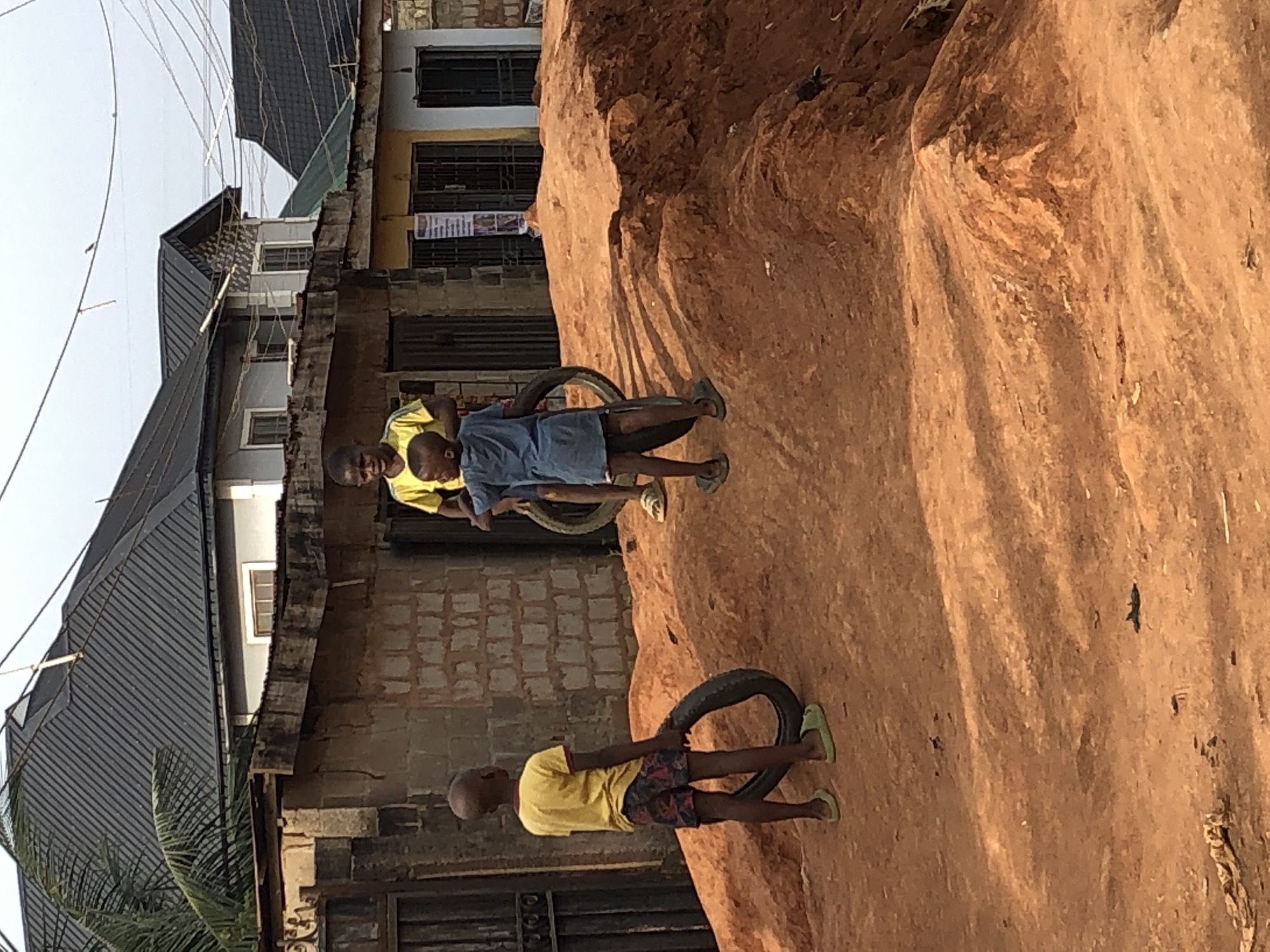 Three boys are playing with old tires in an urban environment. One boy in a yellow shirt and patterned shorts is standing near a tire on the right. Two boys, one in a yellow shirt and the other in a blue shirt, are standing closer to buildings in the background with another tire. The ground is reddish-brown. Buildings are visible behind the boys, including one with a corrugated metal roof. Utility lines stretch across the sky. The location is in the city of Unknown, Unknown.