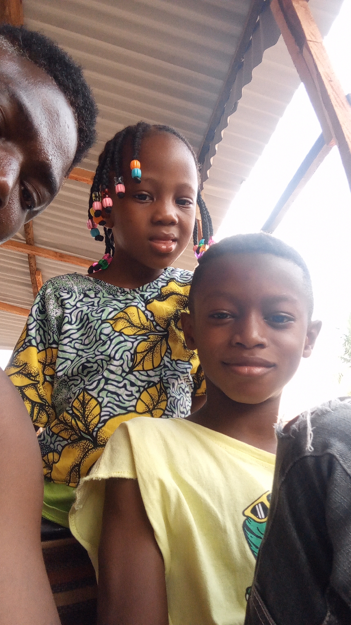 Three individuals are visible in the frame. A dark-skinned individual is partially shown on the left side. A young girl with braided hair adorned with colorful beads is positioned in the upper center, wearing a patterned yellow and green top. A young boy in a yellow t-shirt is in the center-right. The background shows a roof structure with corrugated metal sheets. The setting appears to be outdoors, and the photo may be taken near Sabon Tasha, Nigeria.